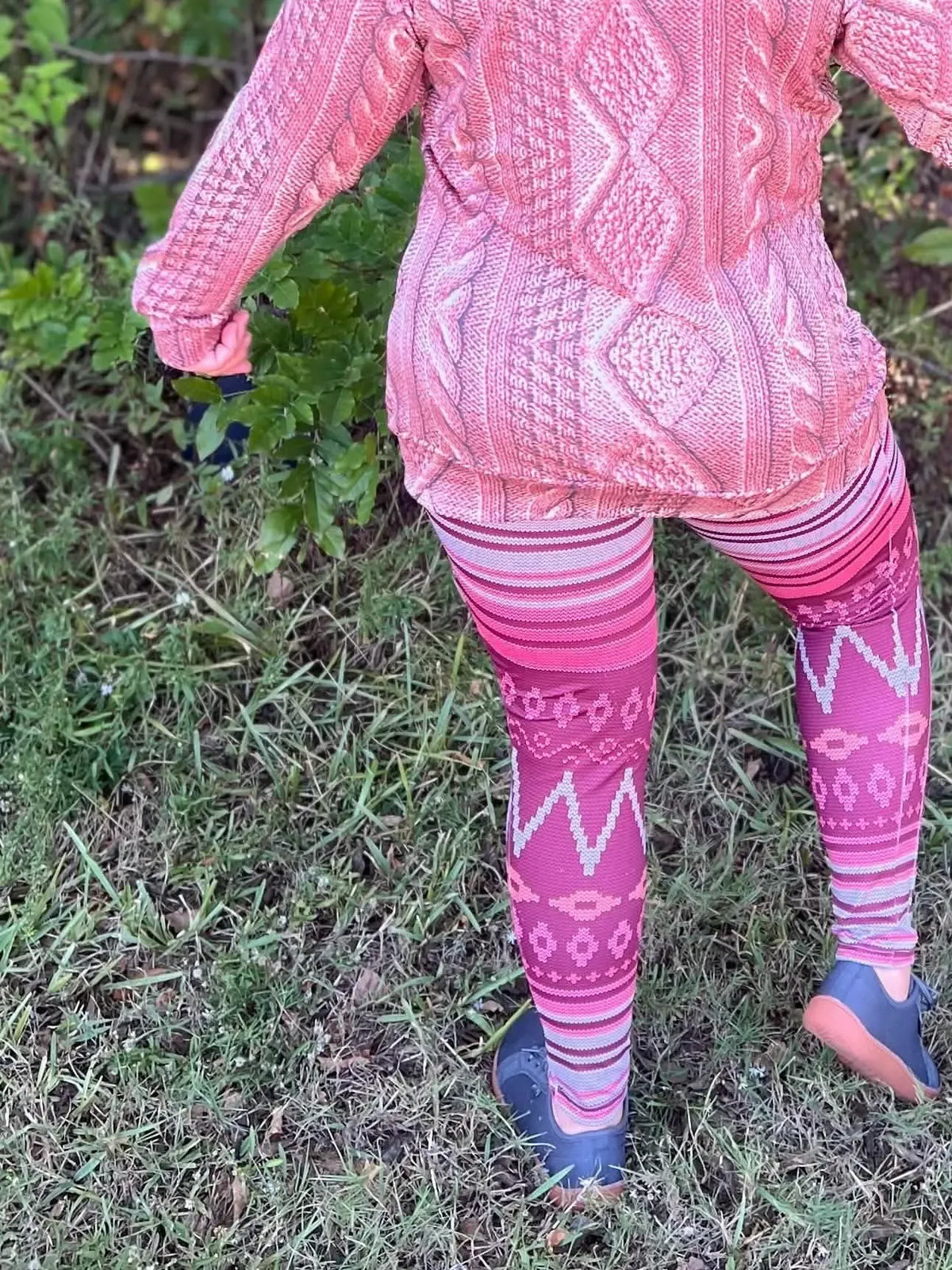 A person in a pink patterned sweater and leggings stands on grass.