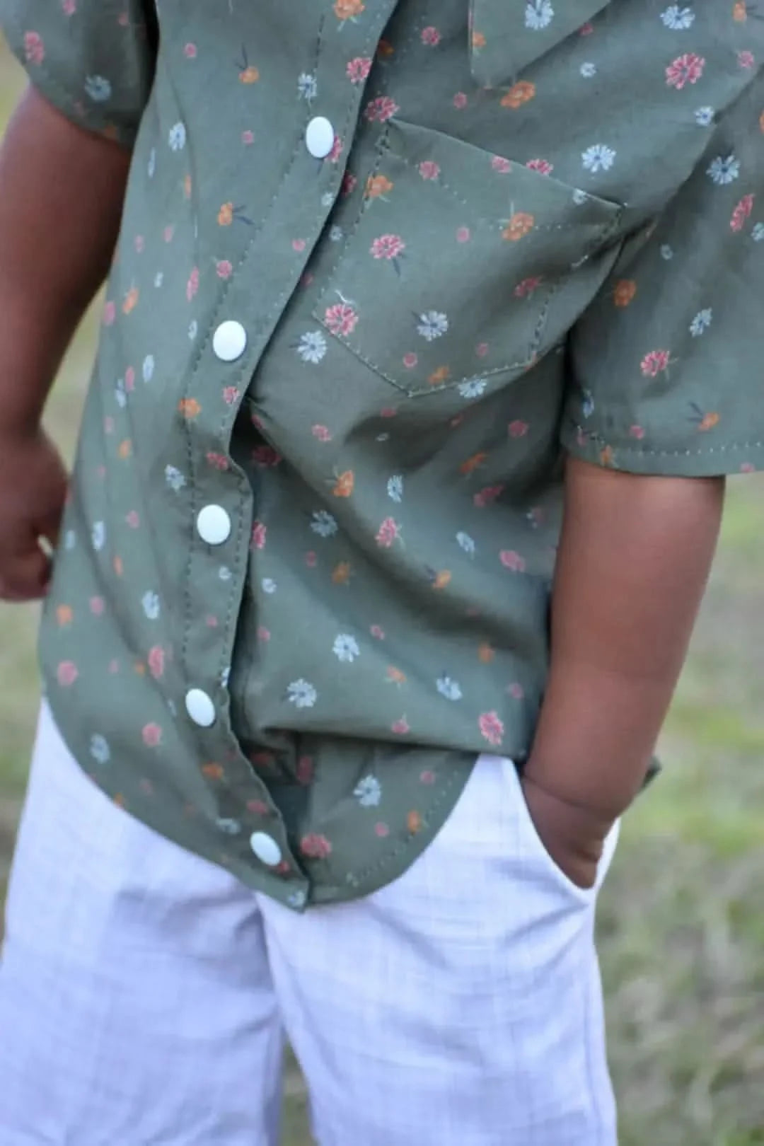 A child wears a green floral shirt with white buttons and light-colored plaid shorts.