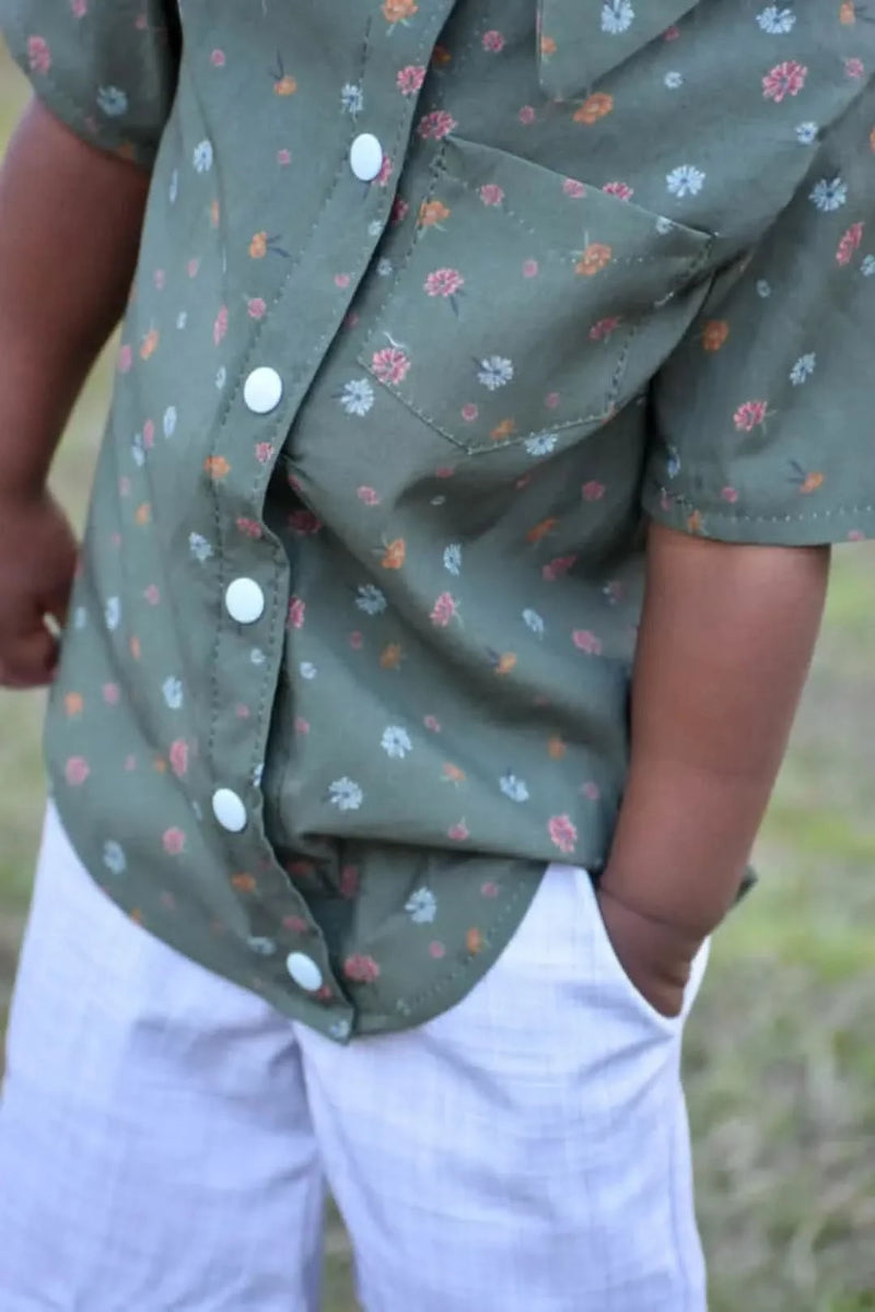 A child wears a green floral shirt with white buttons and light-colored plaid shorts.
