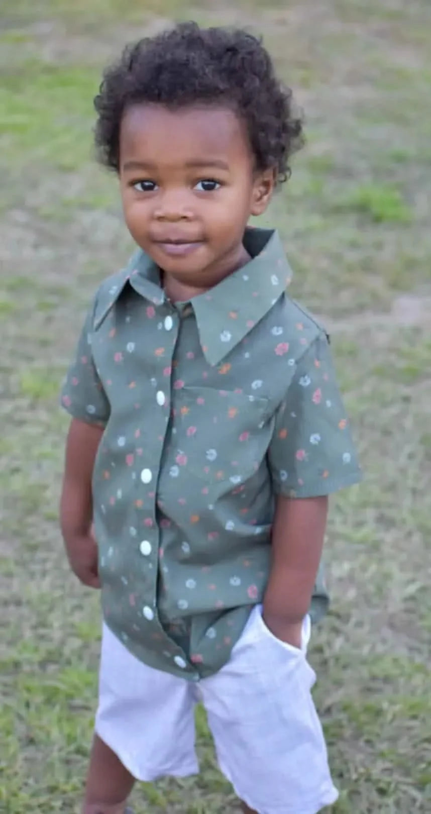 A young child wears a green floral shirt and white shorts.
