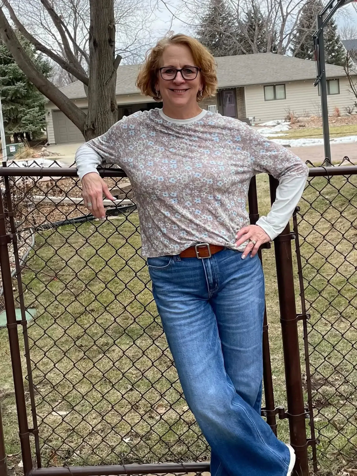Woman in a brown floral top and jeans leaning on a fence.