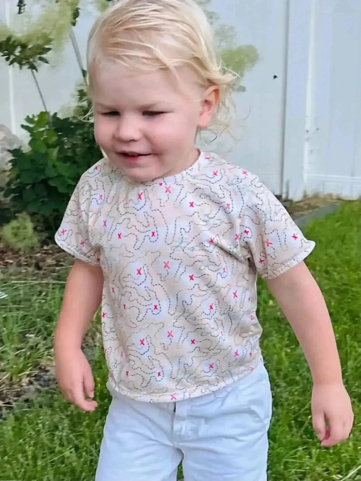 Beige kids' short-sleeve shirt with red pirate adventure print on a white fence background.