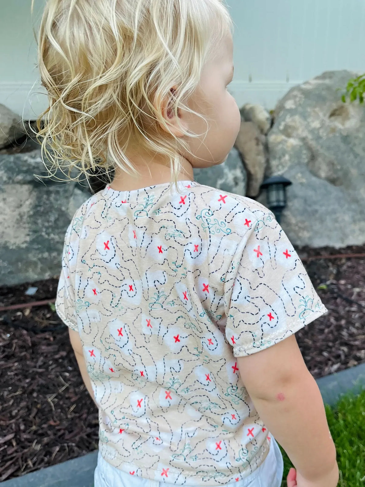 A child from behind wears a beige shirt with a black dashed map and red X's.