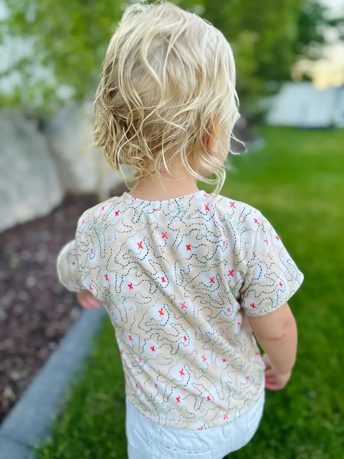 Beige children's short-sleeve shirt with pirate adventure print on grass.