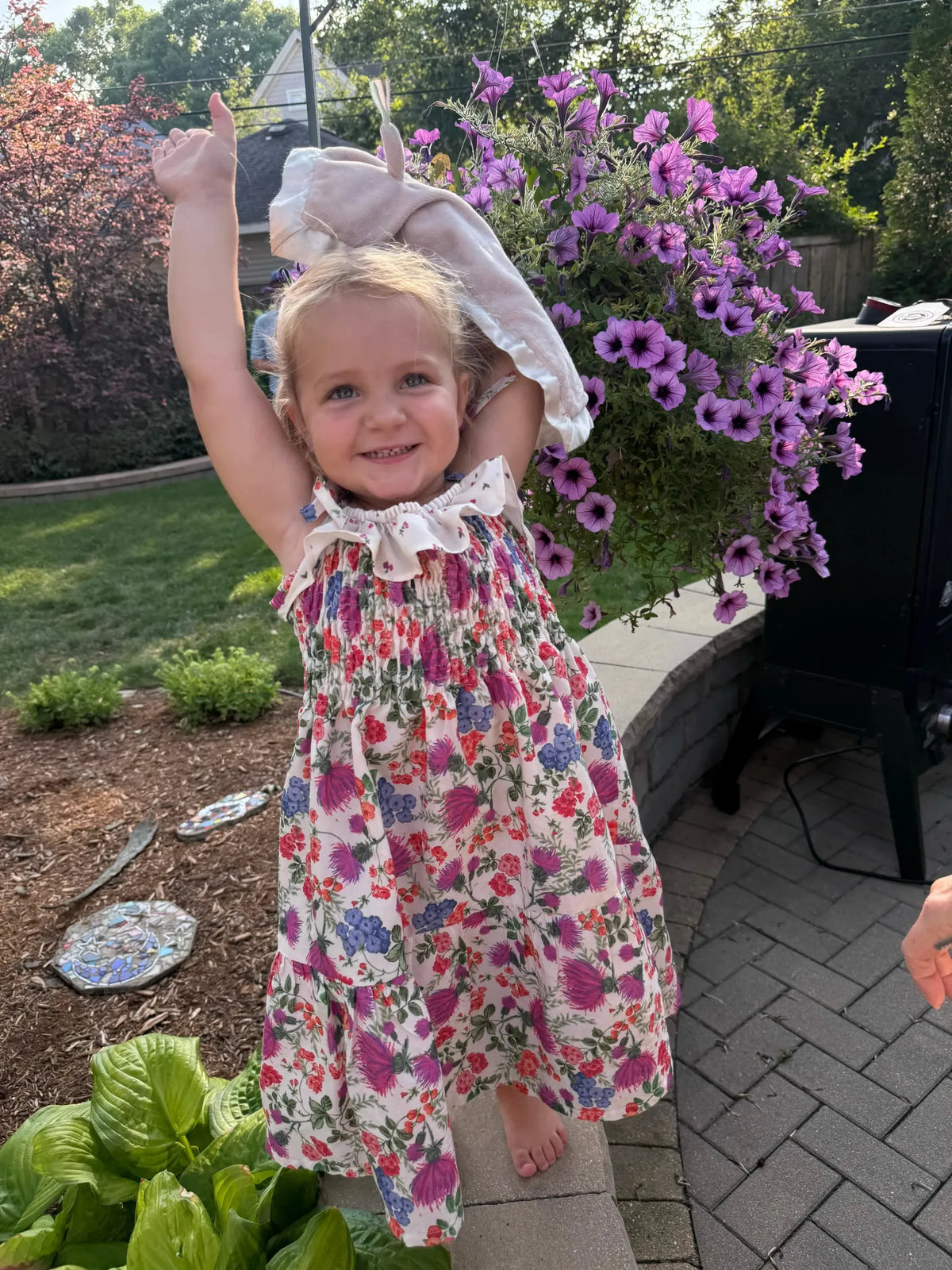 A young girl in a floral dress stands outdoors, holding a hanging basket of purple flowers.
