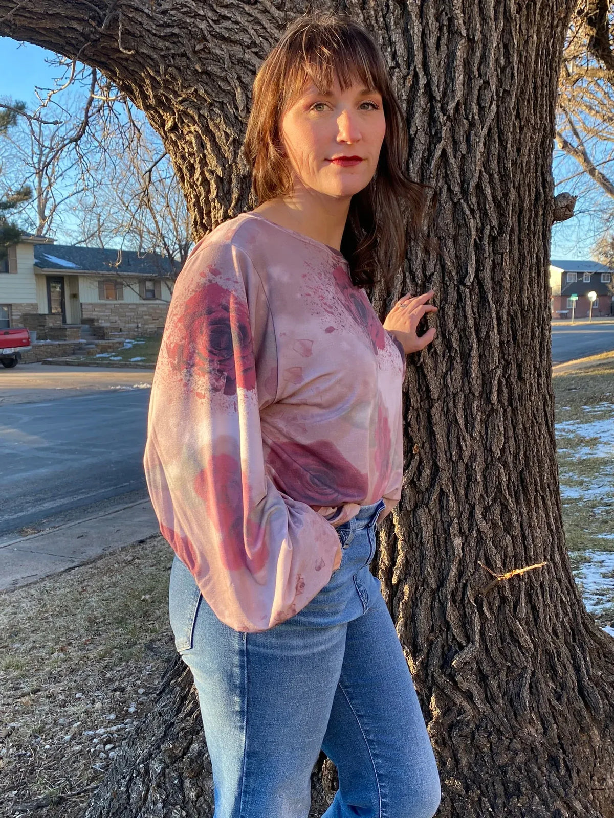A woman wears a pink top with a shattered rose pattern, leaning against a tree.