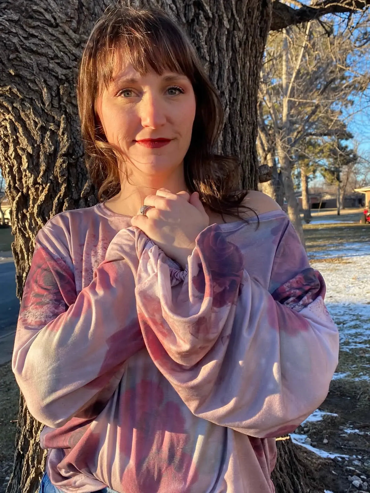 A woman in a pink and purple tie-dye shirt stands by a tree.