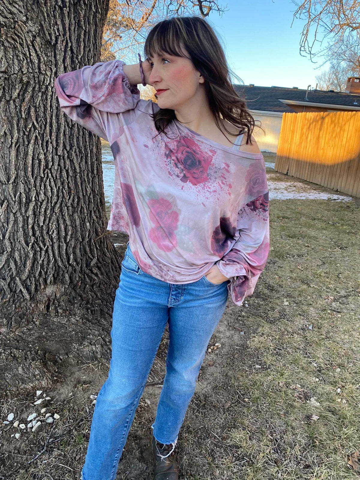 A woman wears a pink and purple floral top with blue jeans, leaning against a tree.