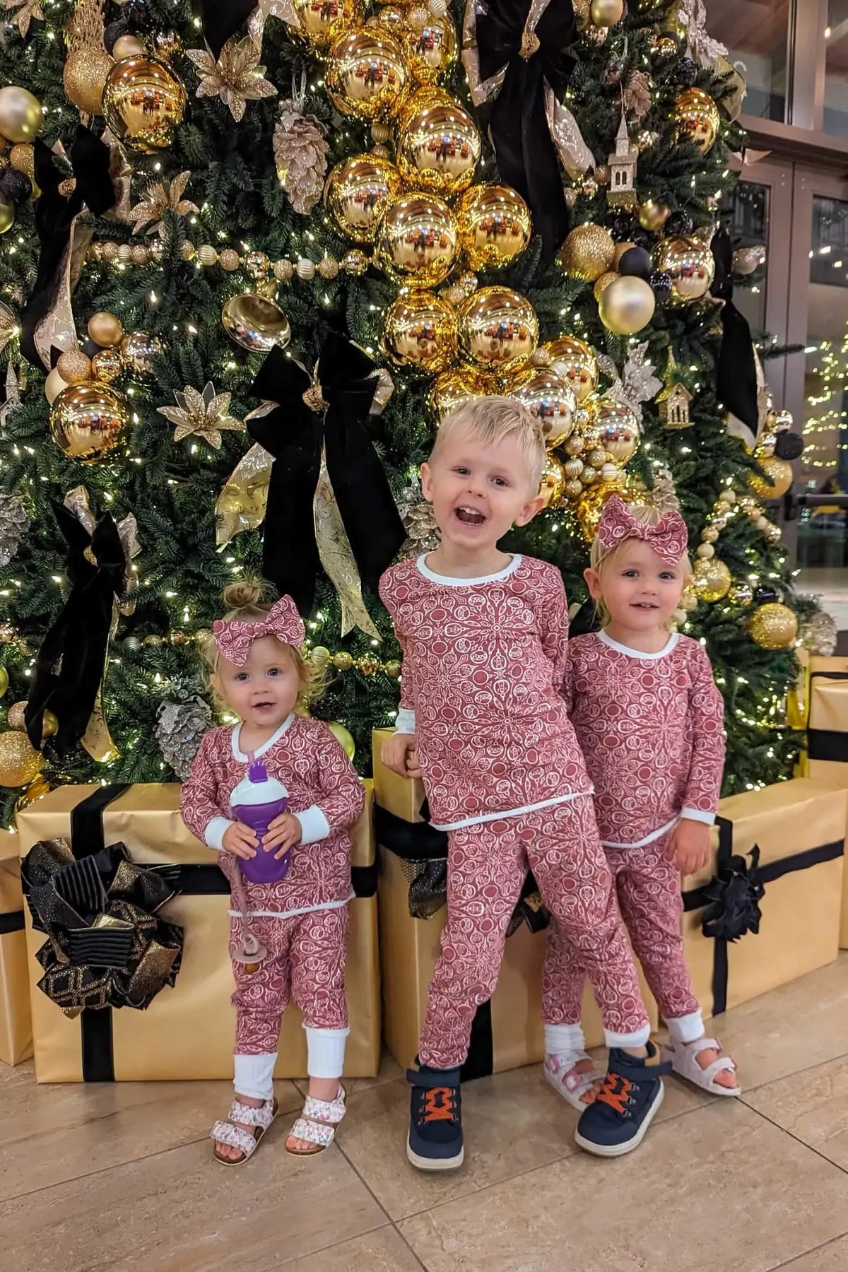 Three children in matching red and white Christmas sweaters stand by a decorated tree.