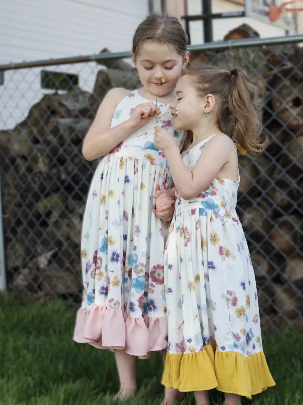 Two girls in floral dresses stand in grass, holding small white flowers.
