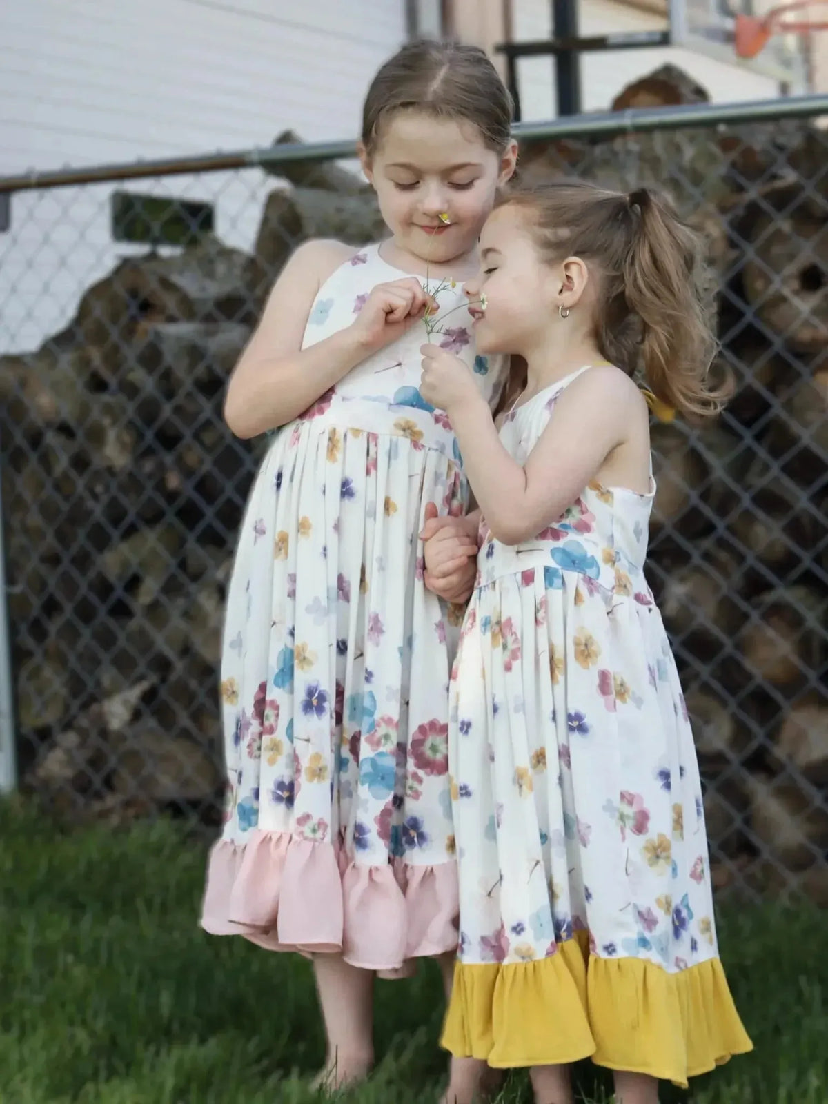 Two girls in floral pastel bloom dresses with ruffled hems against a fence.