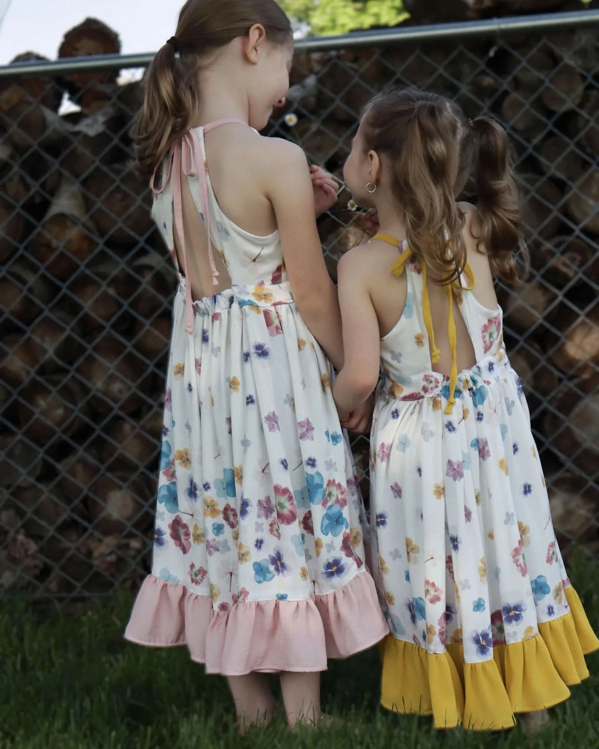 Two girls in floral dresses with ruffled hems stand with their backs to the camera.