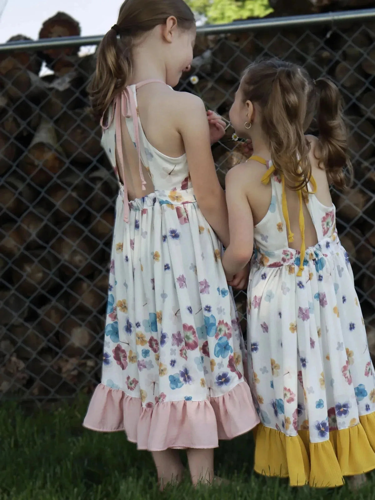 Two girls wear matching white floral sundresses with pastel blooms and ruffled hems.