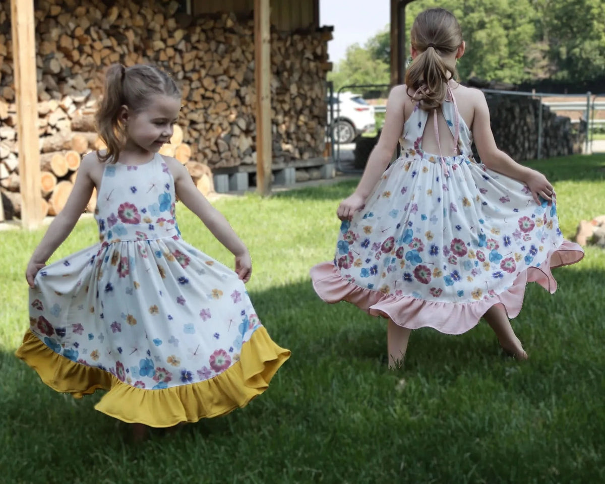 Two young girls in floral dresses twirl in a grassy yard.