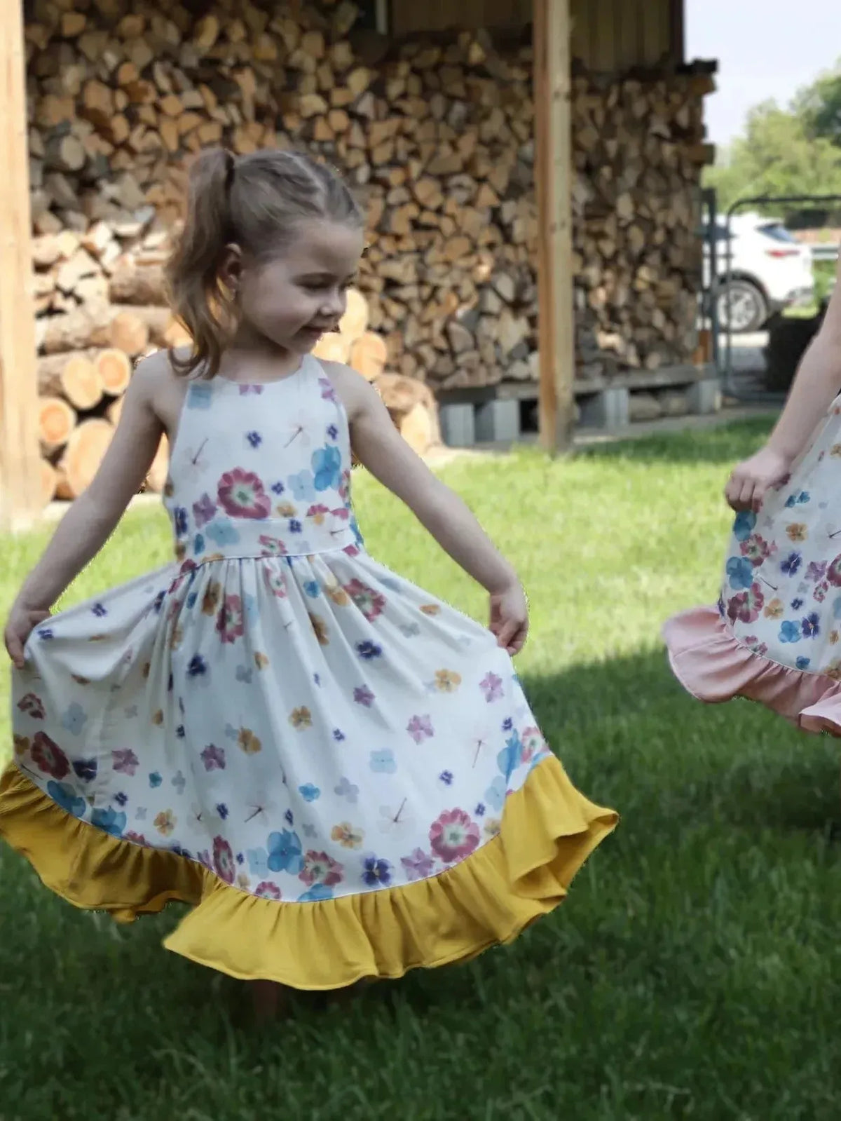 Two young girls twirl in white floral sundresses with yellow ruffled hems on green grass.
