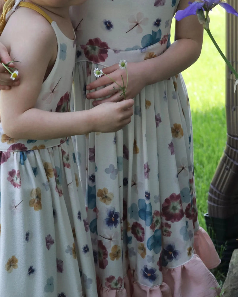 Two children in floral dresses, one holding a daisy, stand in a garden.