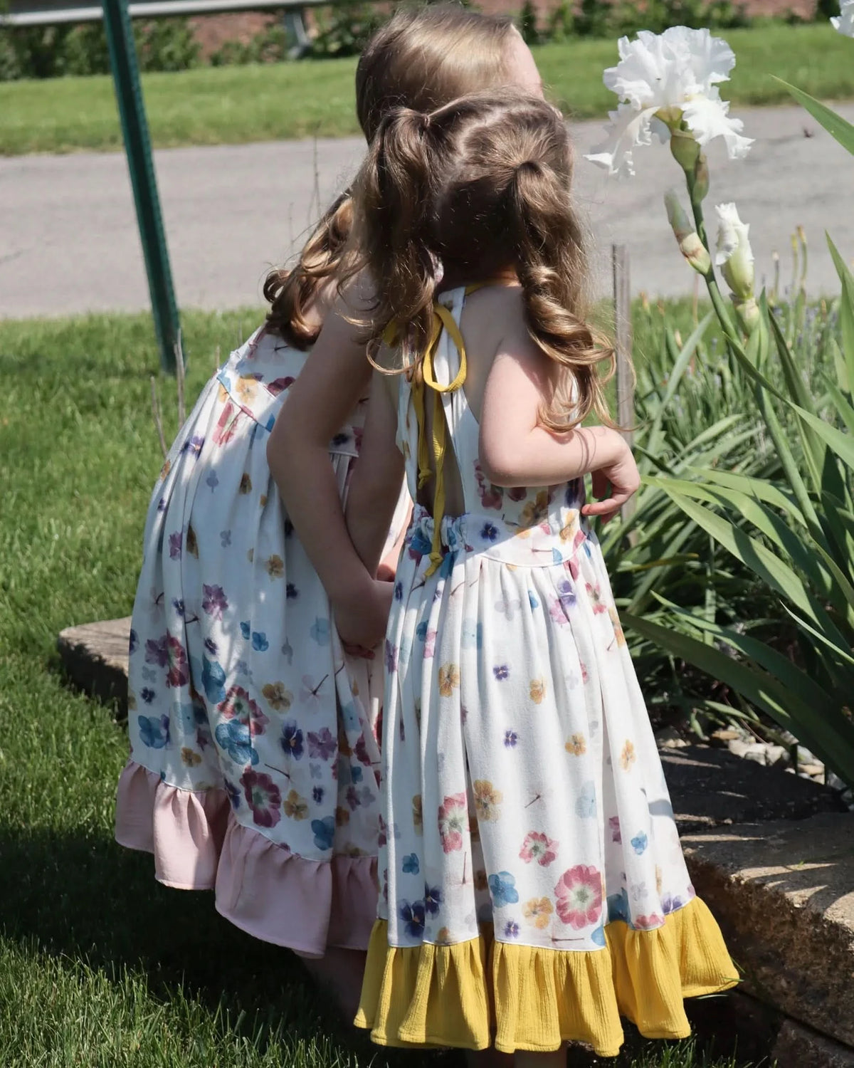 Two girls in floral dresses stand by a white iris in a garden.