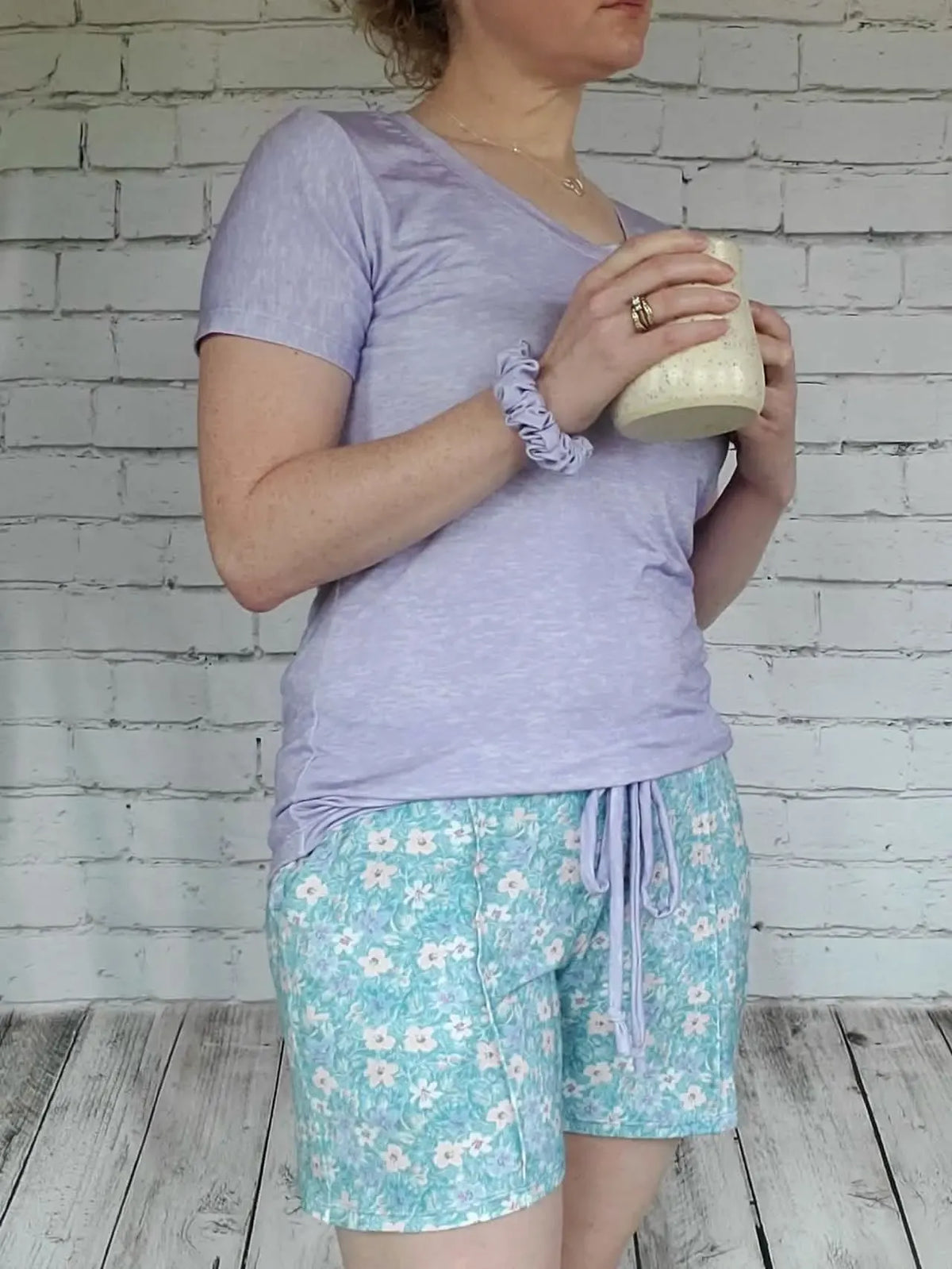A person wears a light purple shirt and floral shorts, holding a mug.