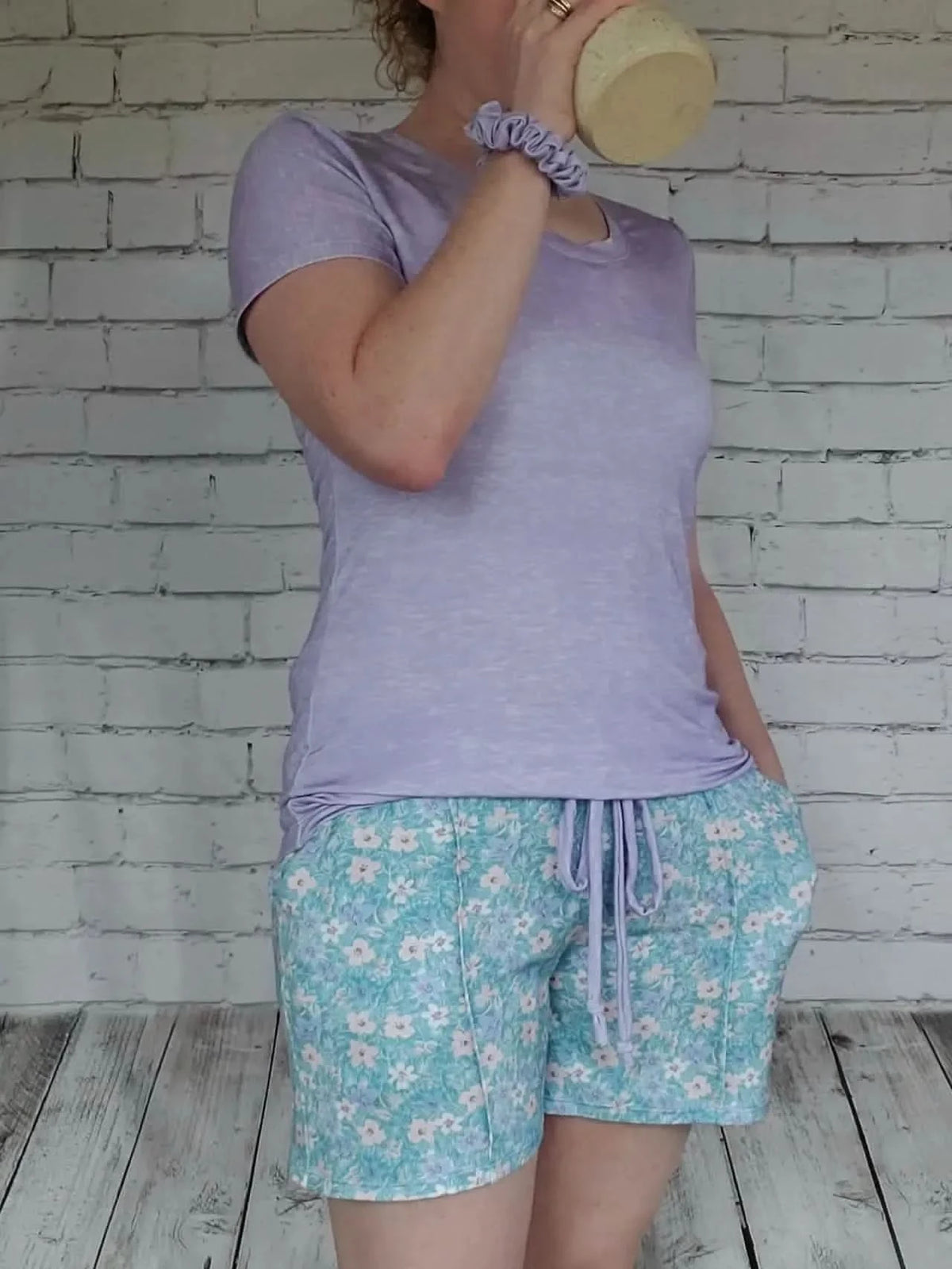 A person wears a lilac heather t-shirt and floral shorts, drinking from a mug.