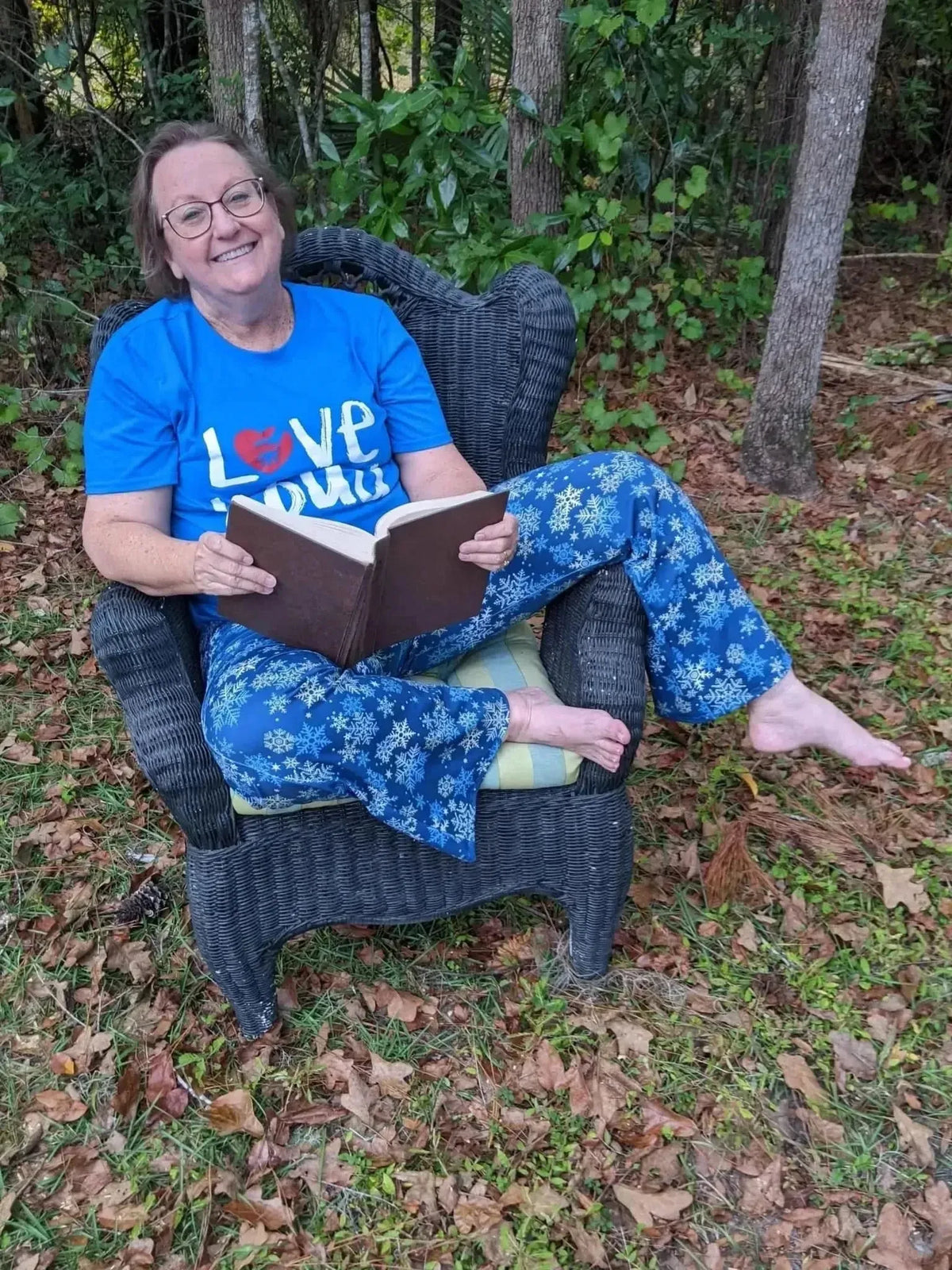 Woman relaxing in chair wearing blue "Love" shirt and snowflake-print pajama pants outdoors.