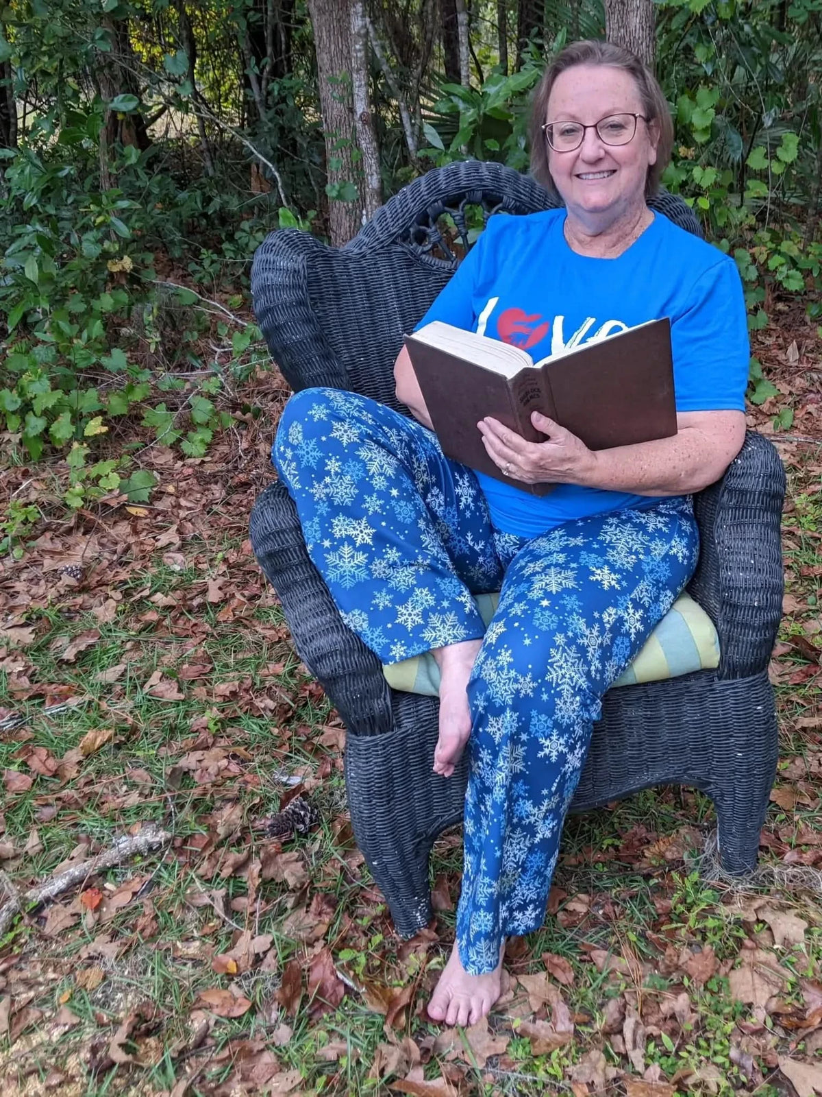 A woman in blue snowflake pajamas sits in a wicker chair, reading a book.