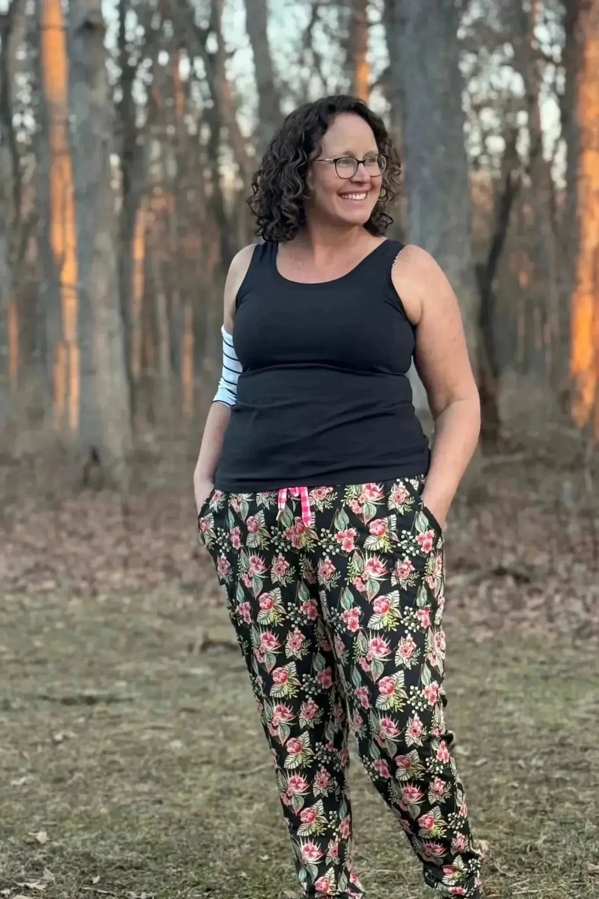 Woman wearing black tank top and floral pants with pink blooms and green leaves in forest.