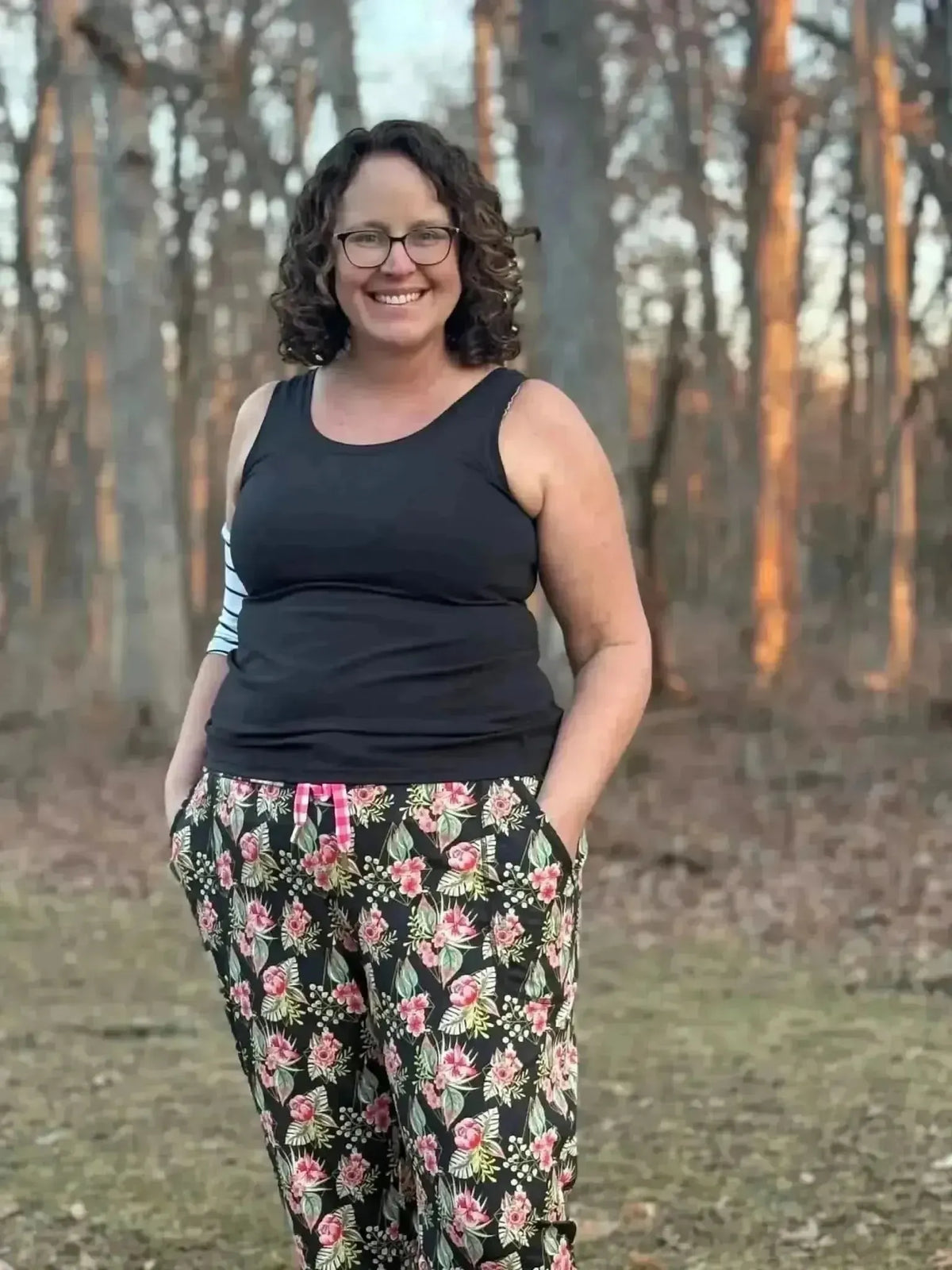 Woman wearing black tank top and pink floral print pants with green leaves in a wooded area.