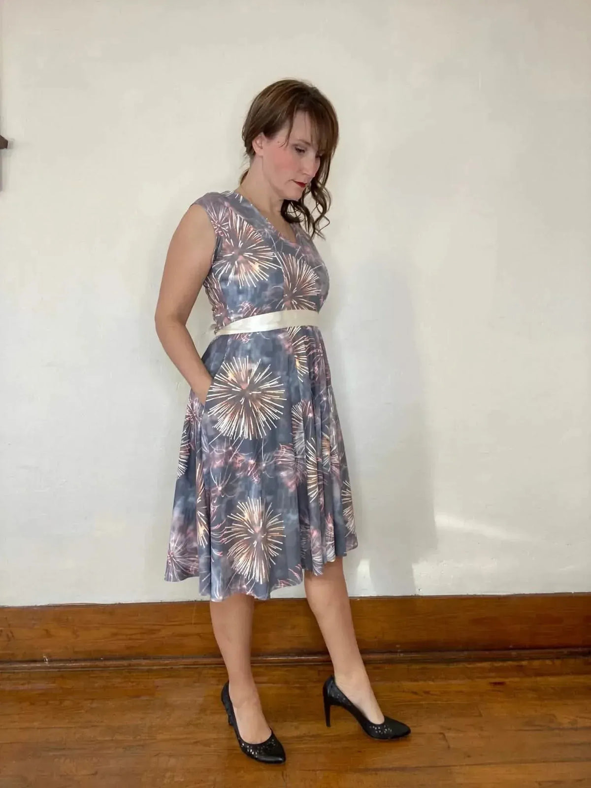Woman modeling a knee-length fit-and-flare dress in gray fireworks print fabric with white sash.