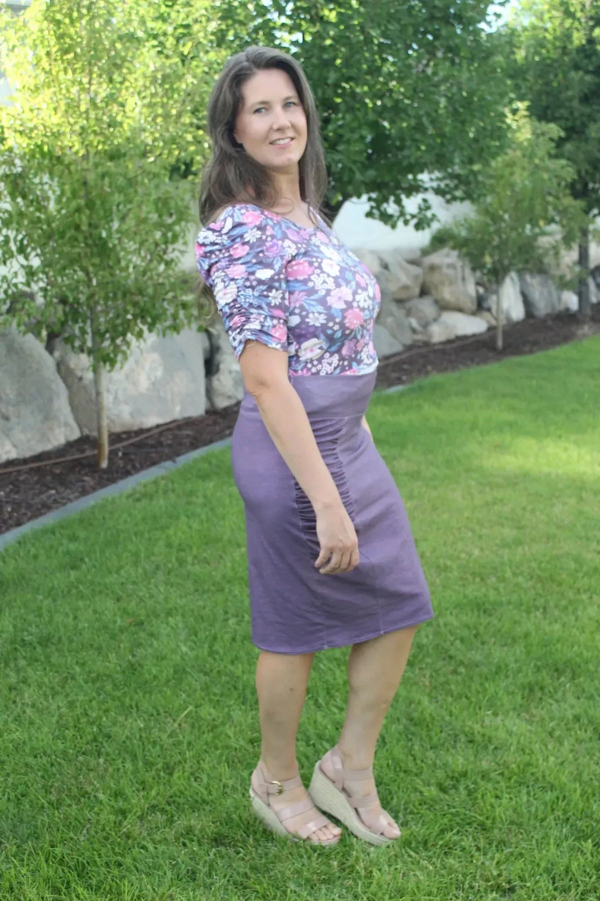 A woman in a floral top and maroon heather skirt stands on grass.