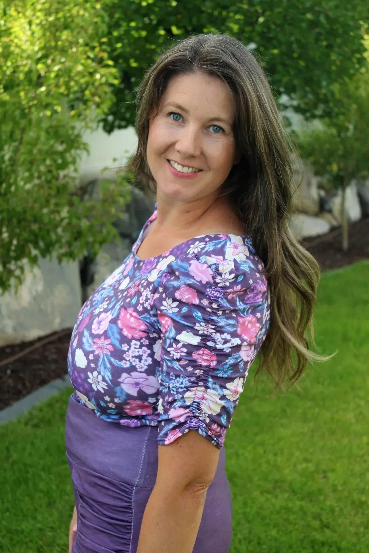 A woman in a purple floral top and skirt smiles outdoors.