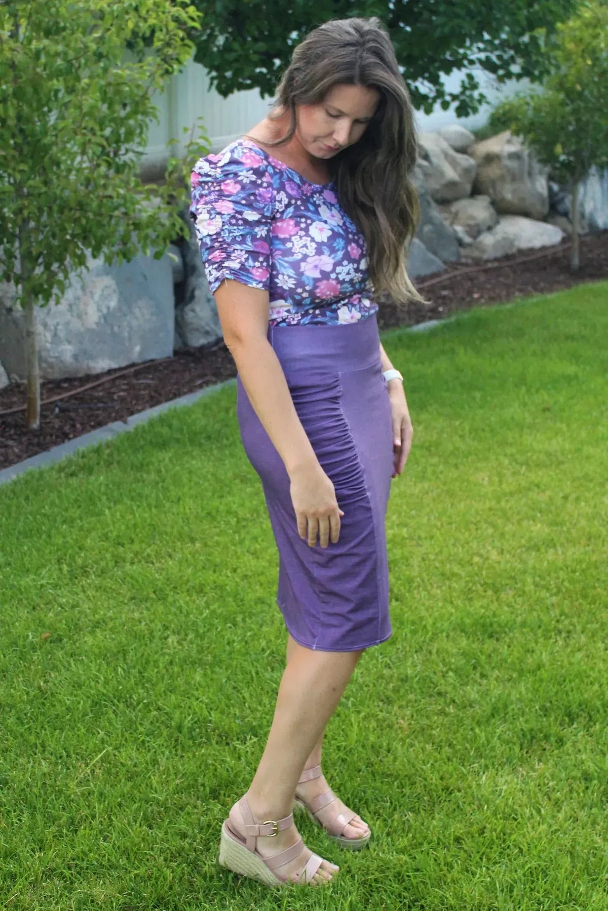 A woman wears a purple floral top and a ruched purple skirt with wedge sandals.