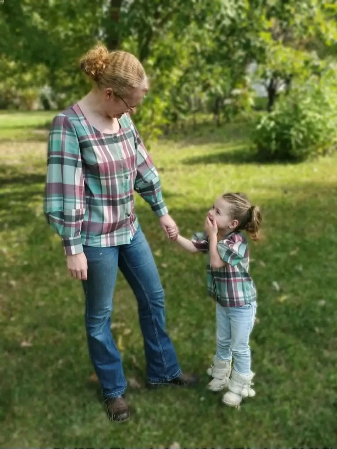 A woman and child wear matching plaid shirts in a green outdoor setting.