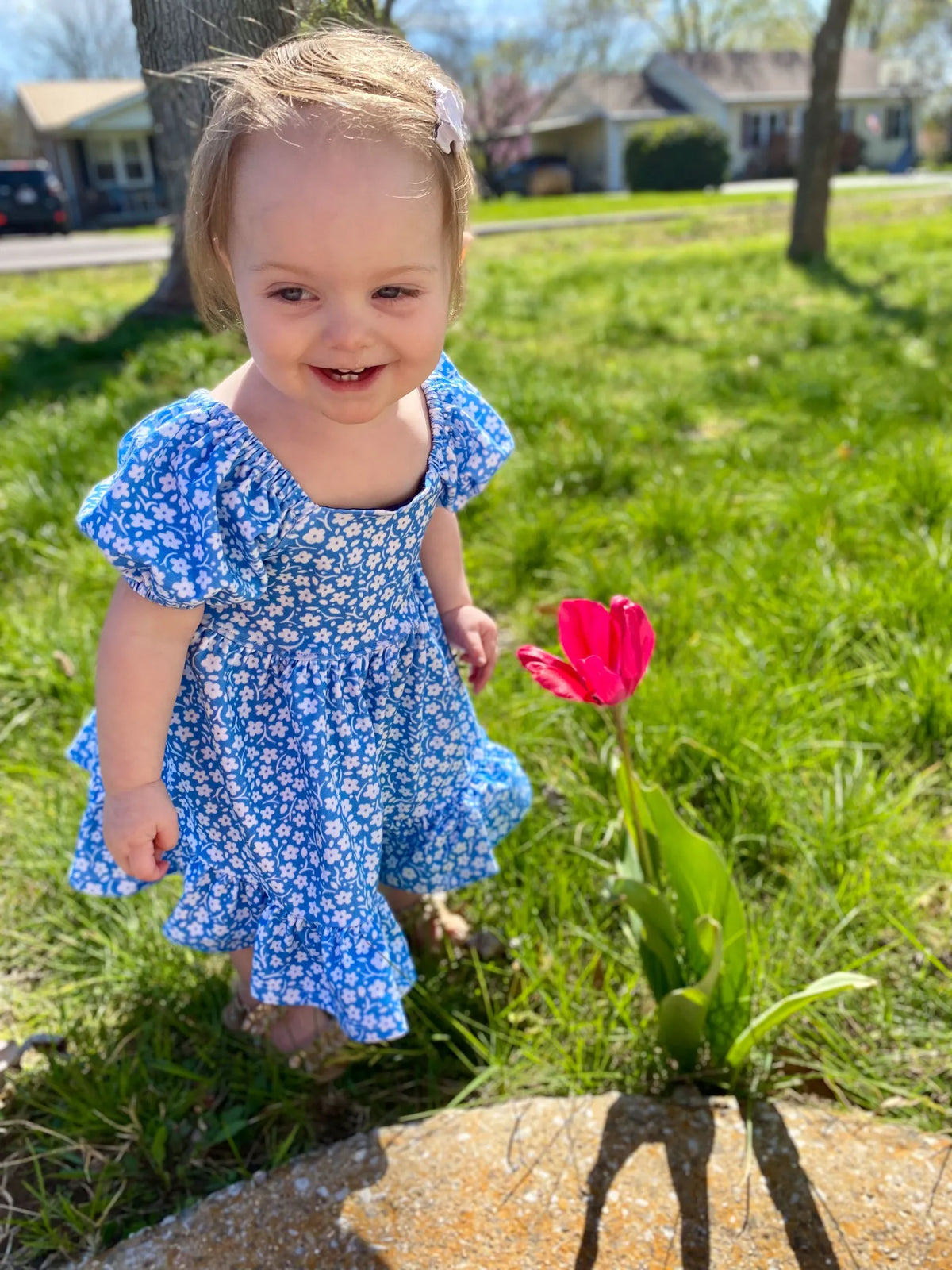 A smiling toddler in a blue floral dress stands next to a pink tulip.