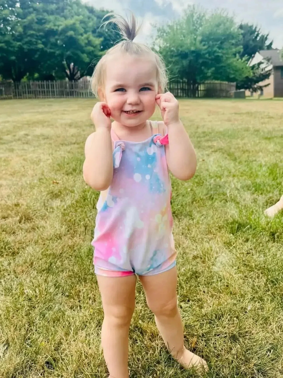 Blonde toddler girl smiling in a pink and purple sparkle print cotton romper on grass.