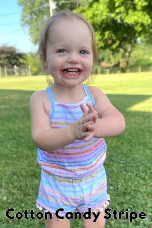A smiling toddler in a cotton candy stripe romper with hands clasped.