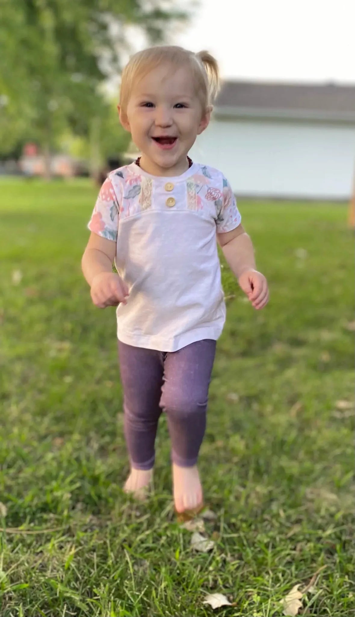 A smiling toddler in a cream heather shirt with floral sleeves and purple pants.