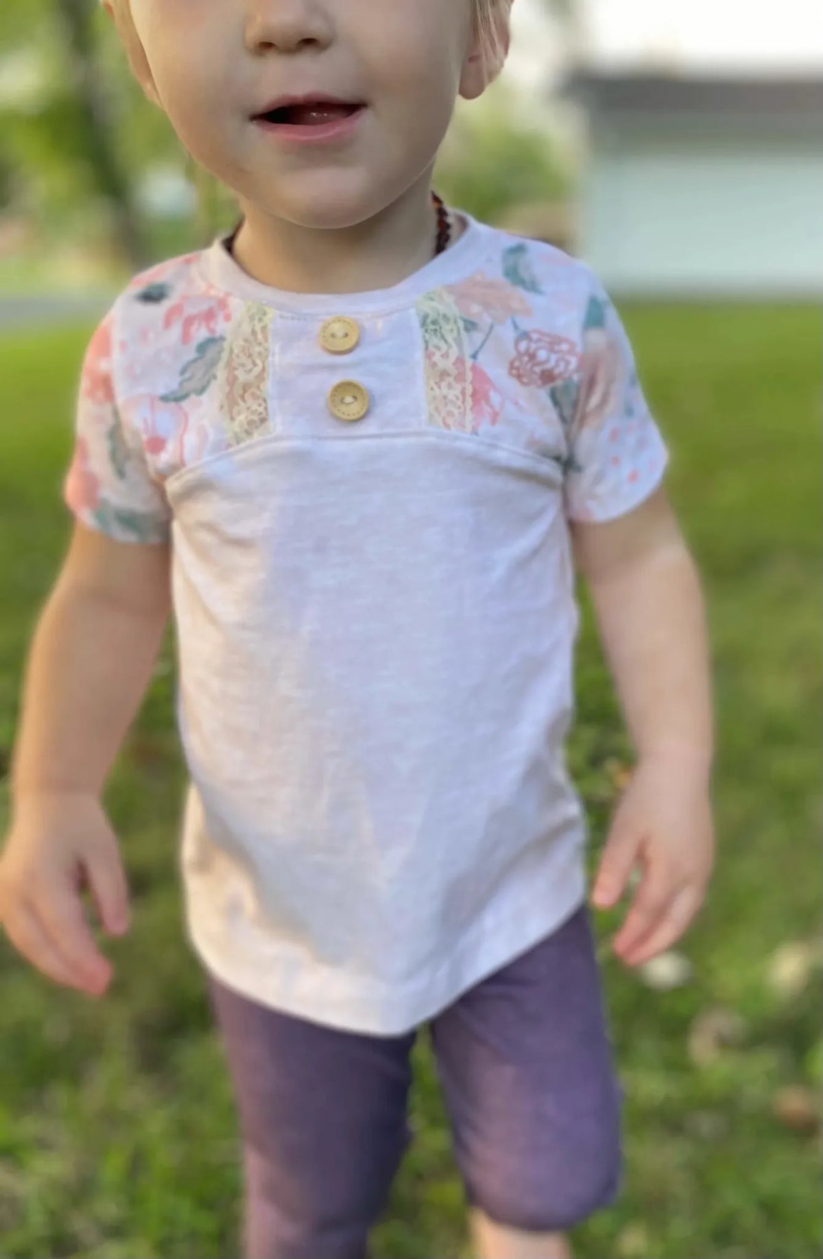 A child wears a cream heather shirt with floral sleeves and purple pants.