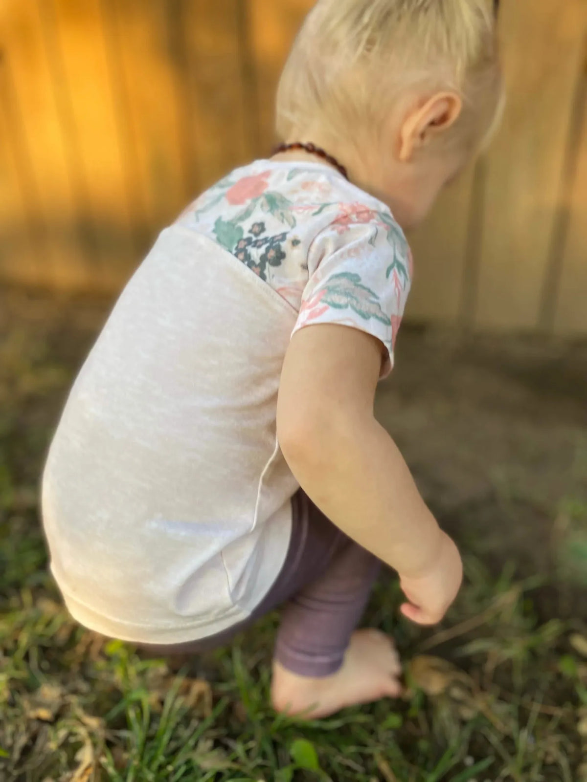 A child in a cream heather shirt with floral sleeves squats on grass.