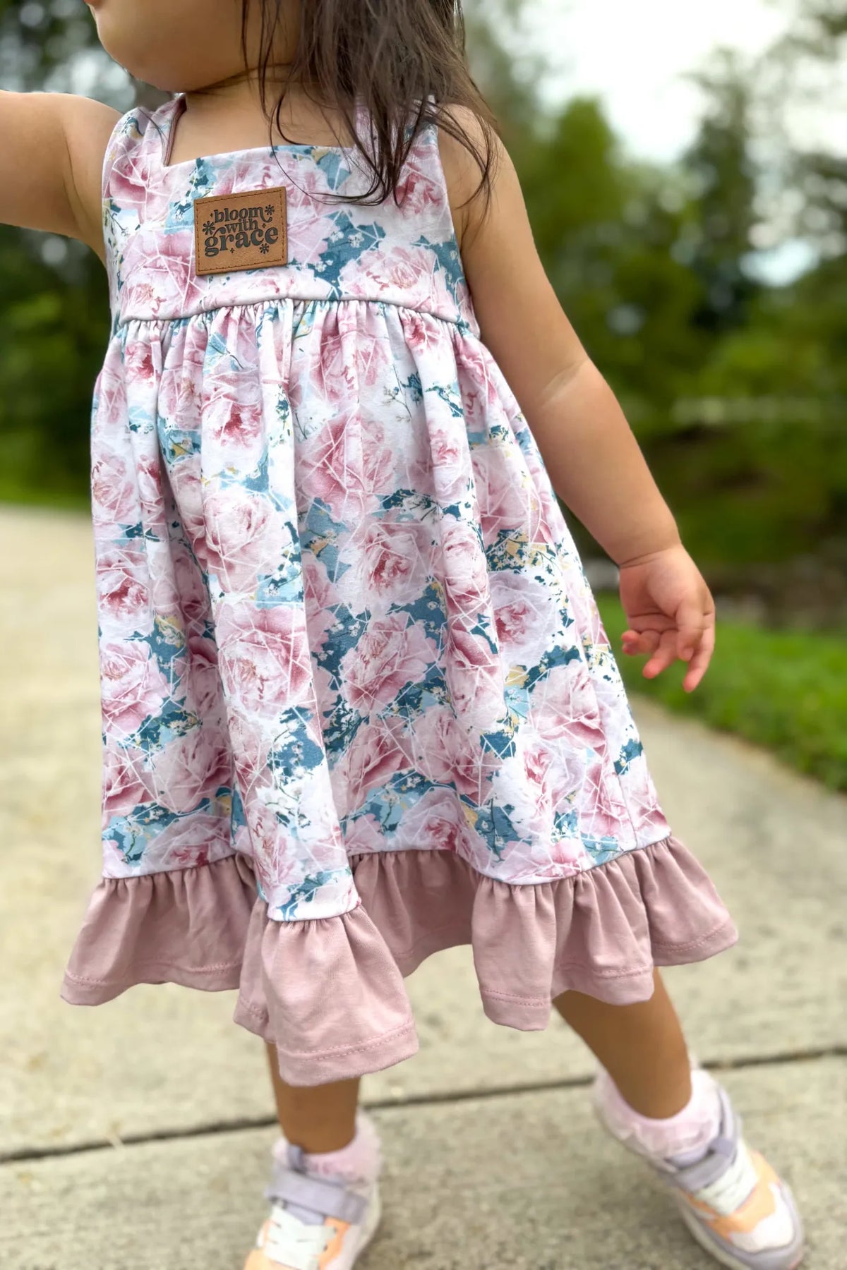 A child wears a dress with a pink and blue floral pattern and a "bloom with grace" tag.