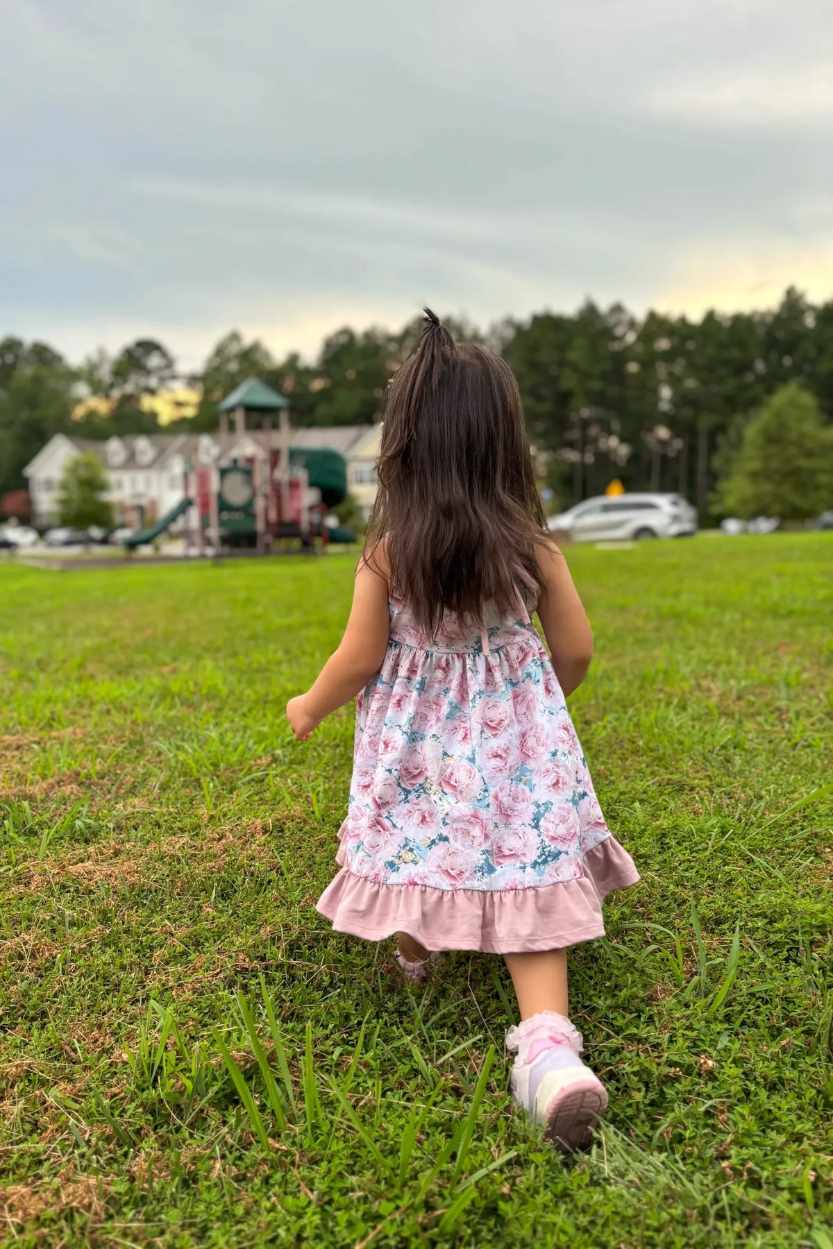 A young girl in a floral dress walks across a grassy field towards a playground.