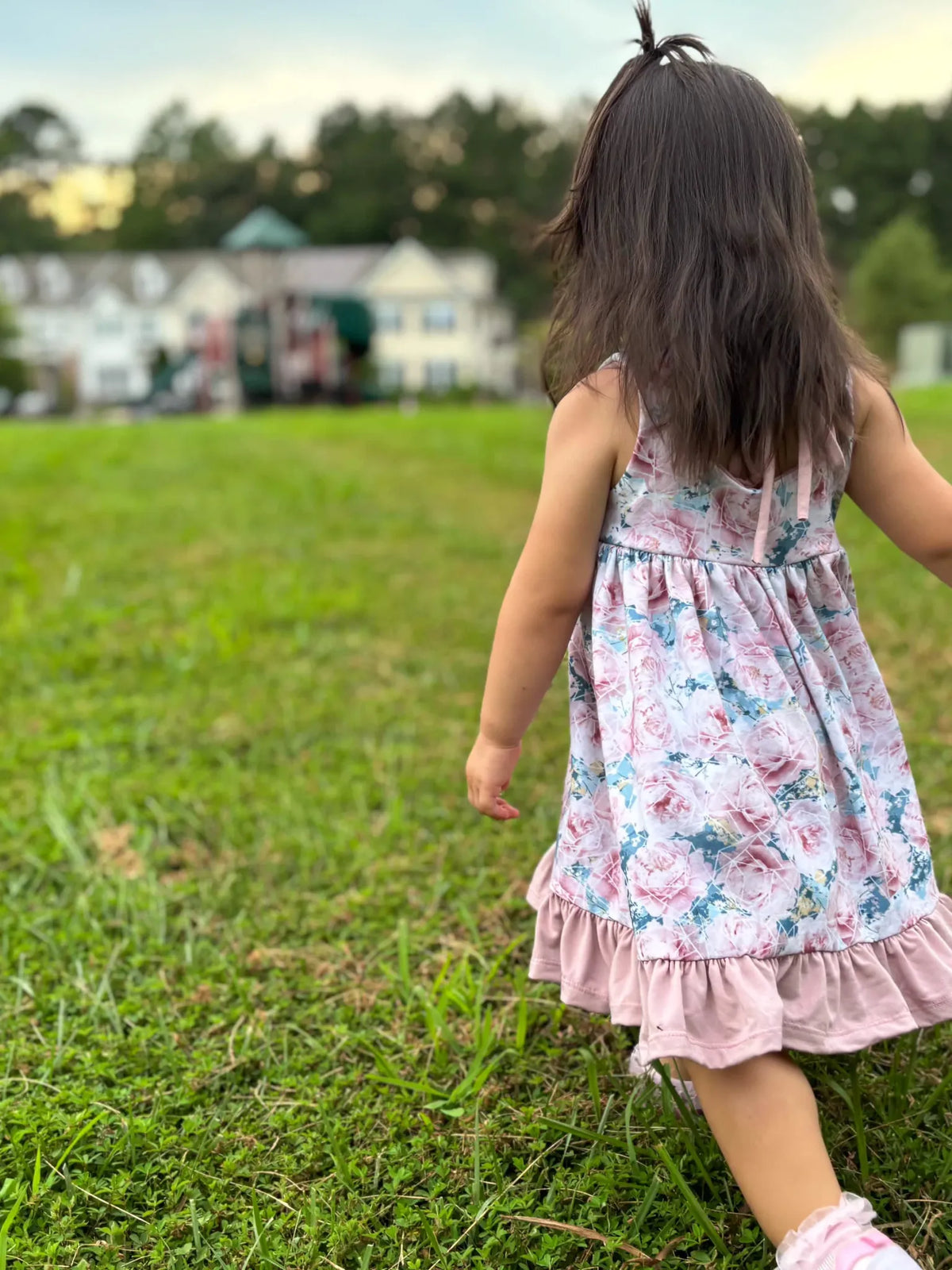 A young girl in a floral dress walks across a grassy lawn.