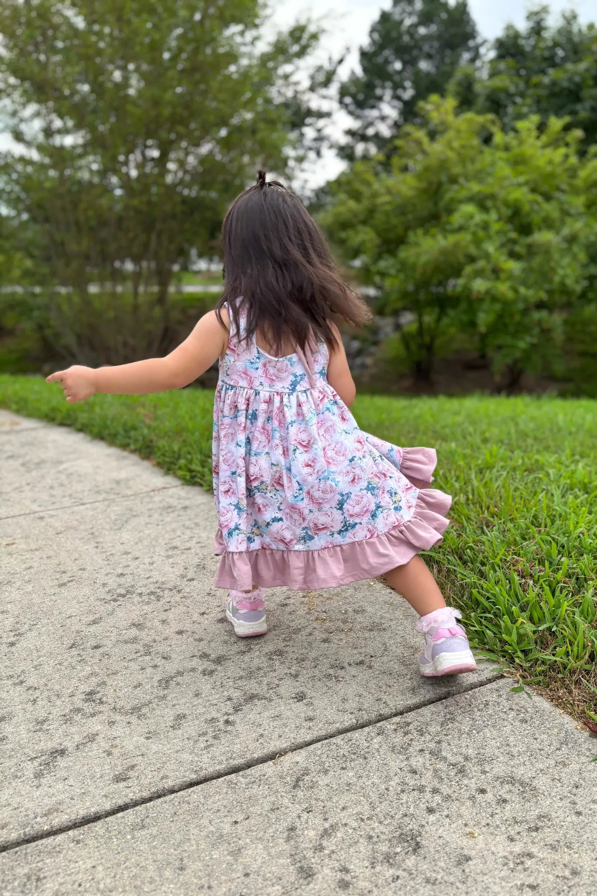 A young girl in a floral dress with a ruffled hem points to the left on a sidewalk.