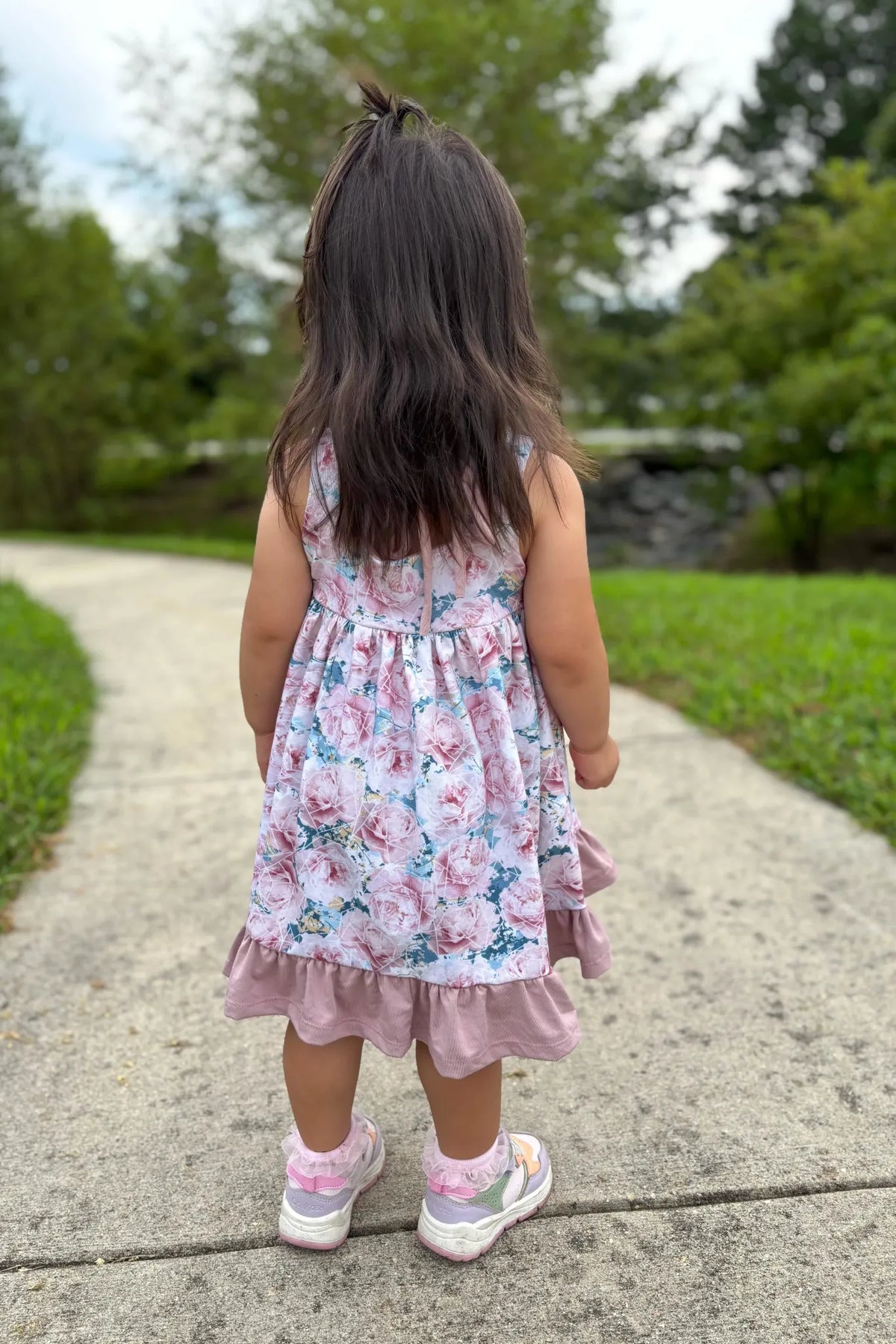 A young child in a floral dress stands on a path with their back to the camera.