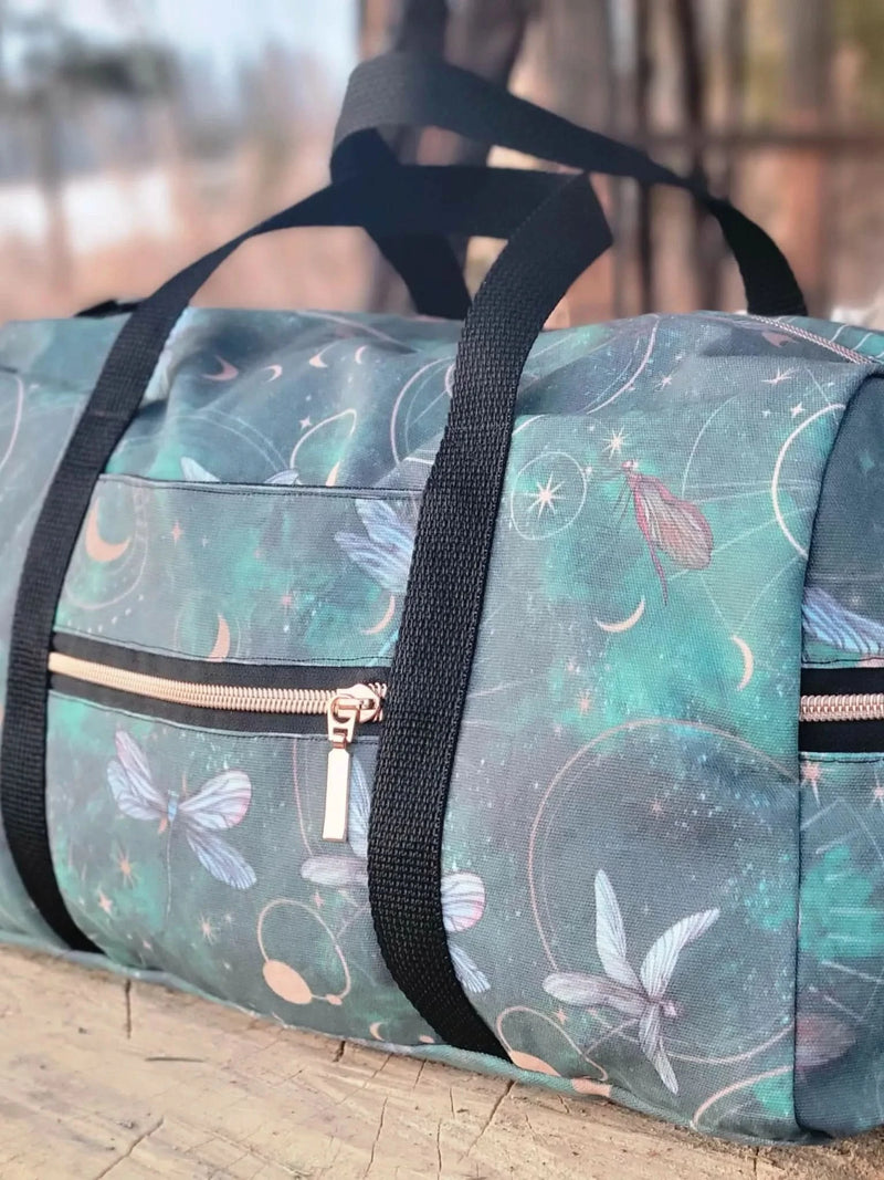 A duffel bag with a celestial dragonfly pattern in green, black straps, and rose gold zippers.