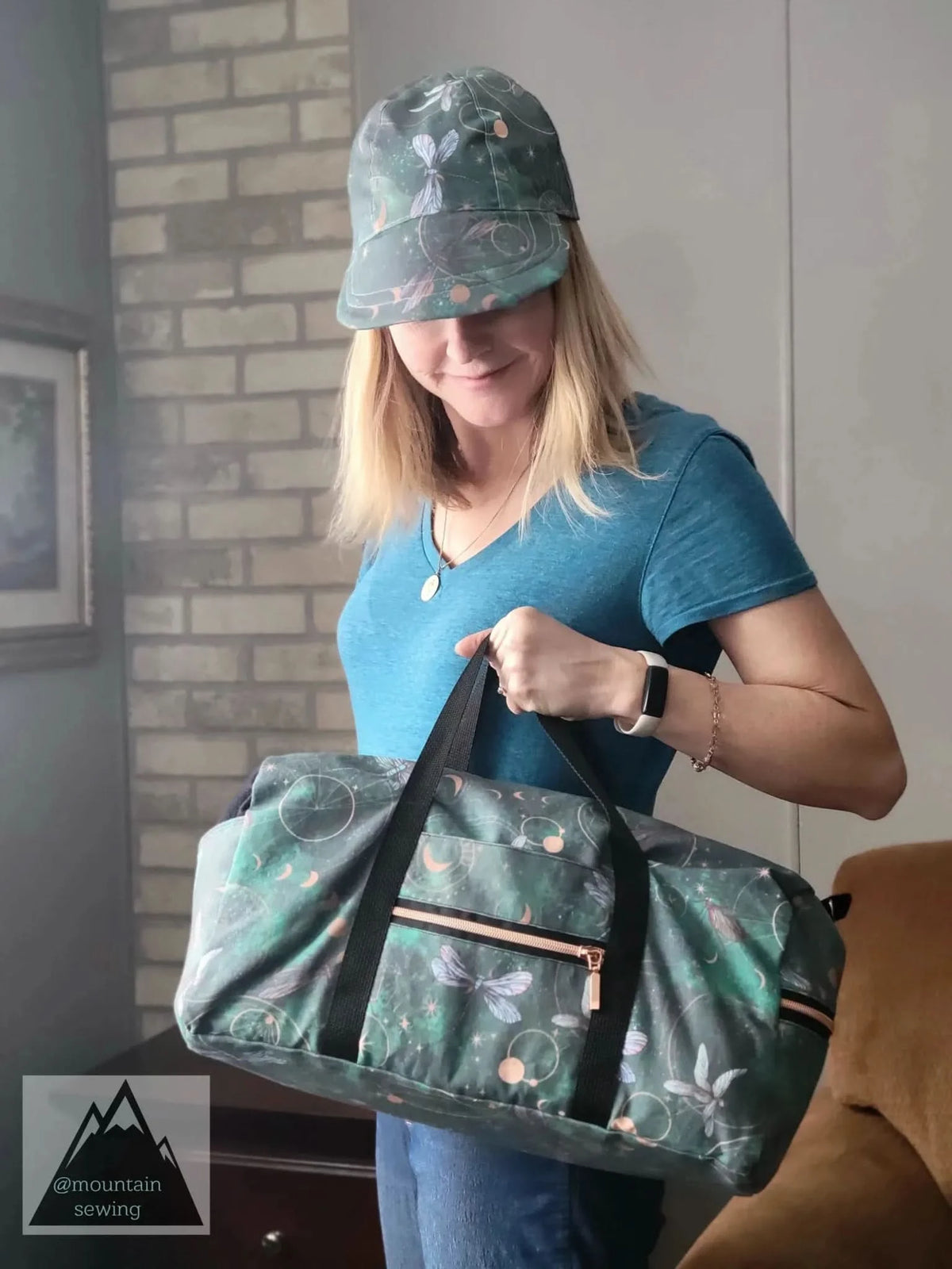 A woman holds a green duffel bag and wears a matching cap with celestial dragonfly print.