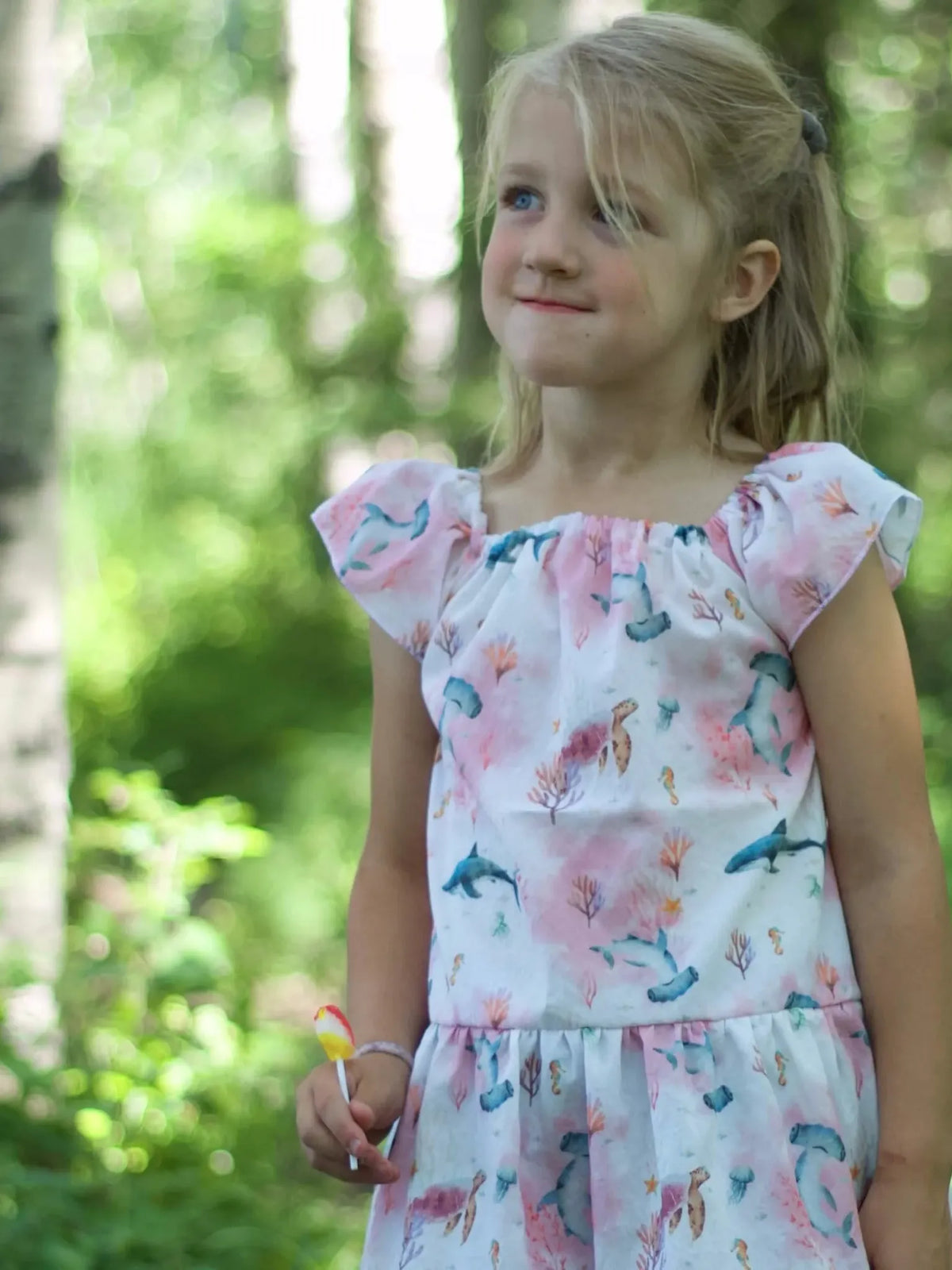 A young girl in a pink "Under The Sea" dress holds a lollipop in a forest.