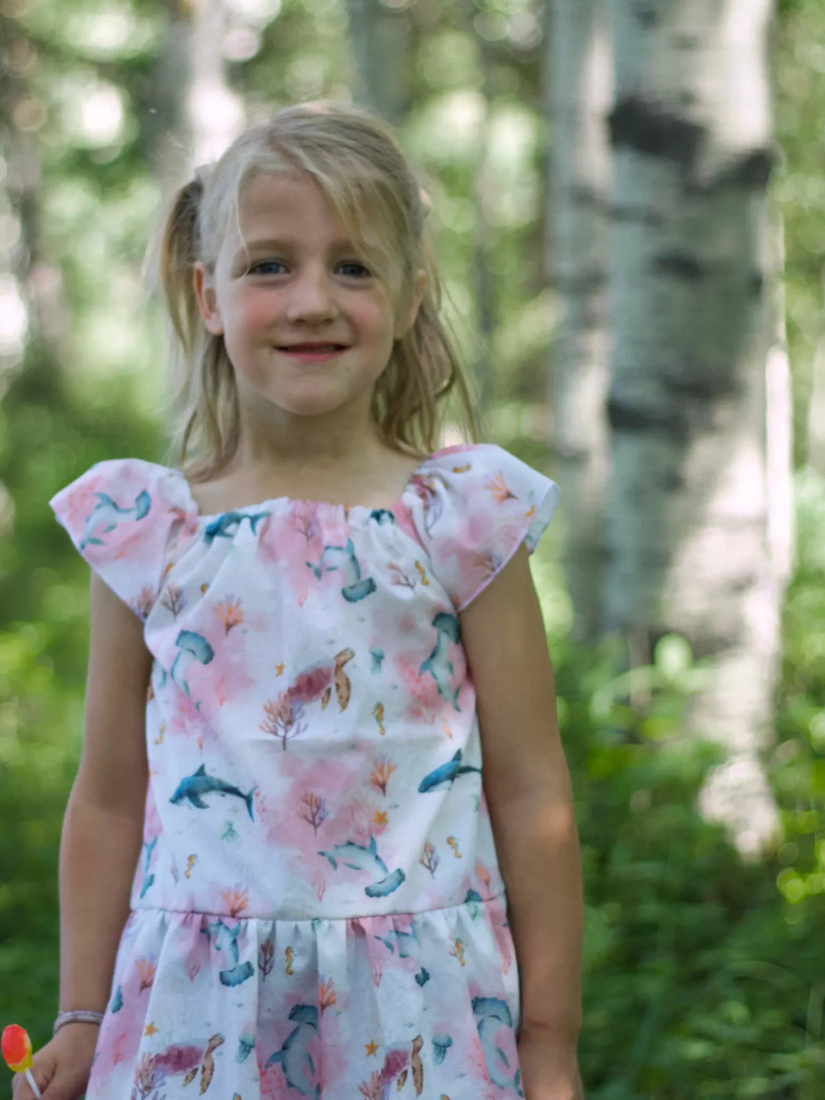 A young girl in a pink dress with an "Under The Sea" pattern stands in a forest.