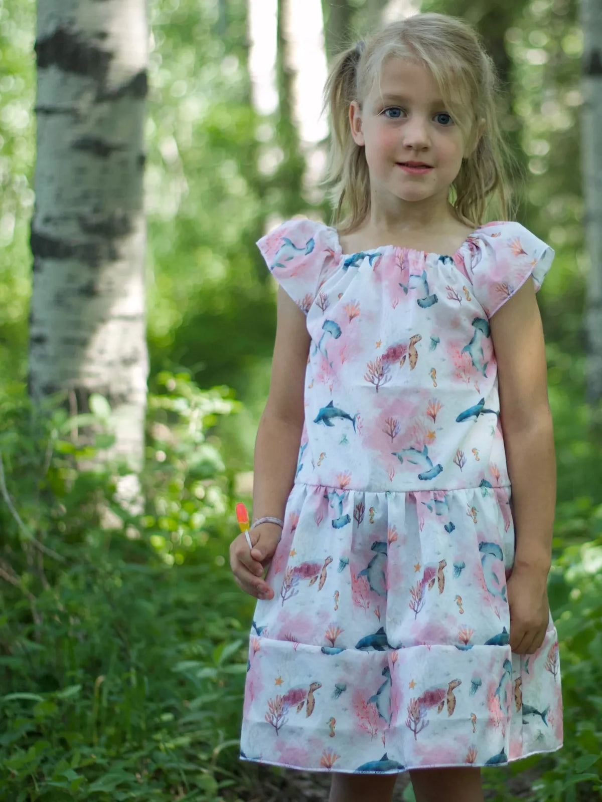 A young girl wears a pink "Under The Sea" dress in a forest setting.