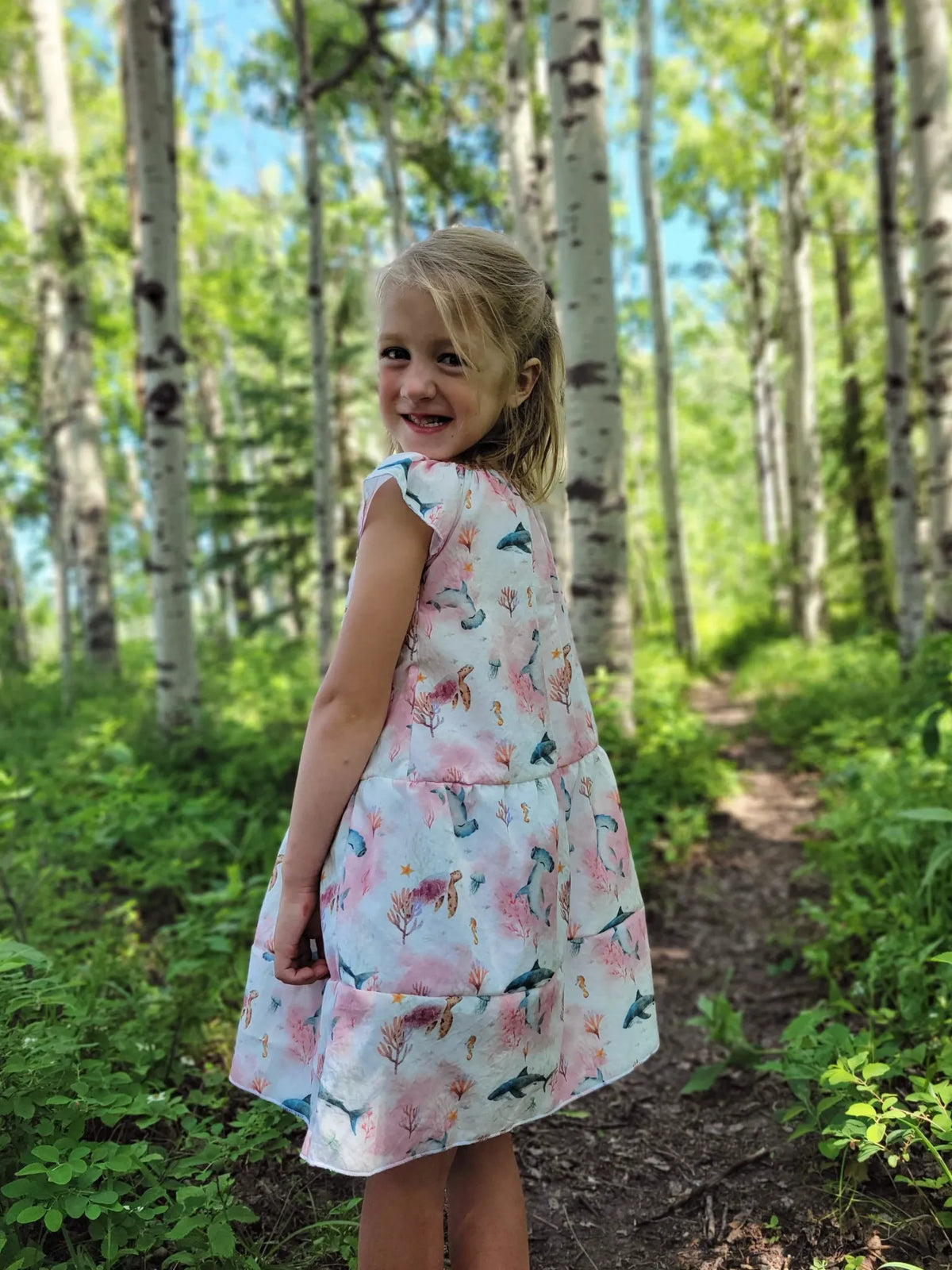 A young girl in a pink "Under The Sea" dress smiles at the camera in a forest.