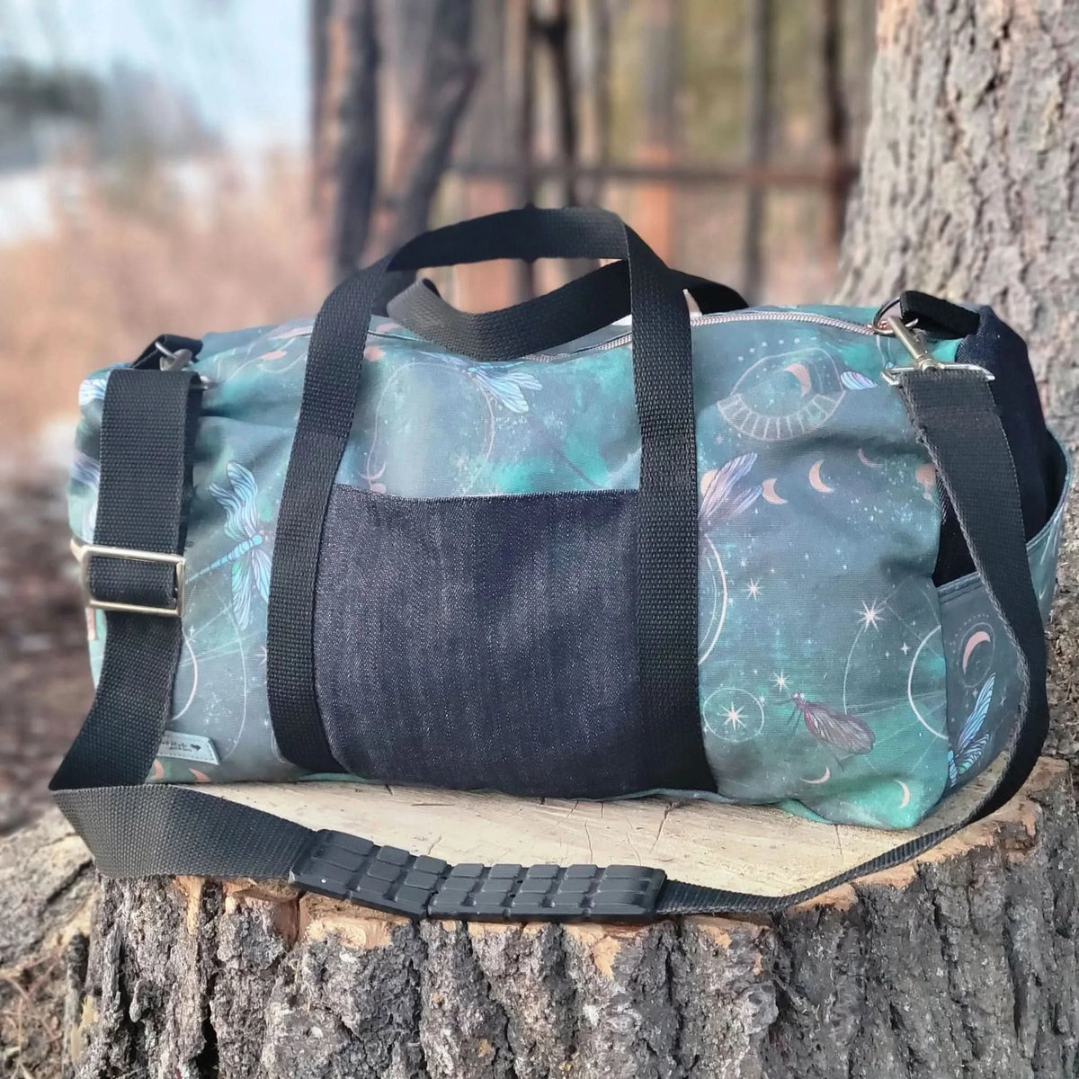A green duffel bag with celestial dragonfly print and black straps rests on a tree stump.